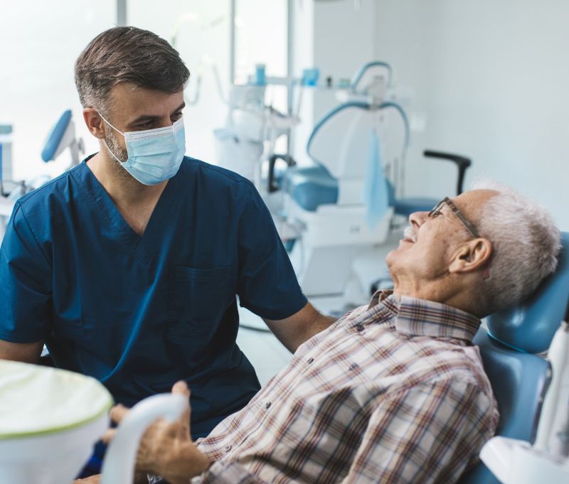 A dentist wearing a mask is talking to an older man in a dental chair.