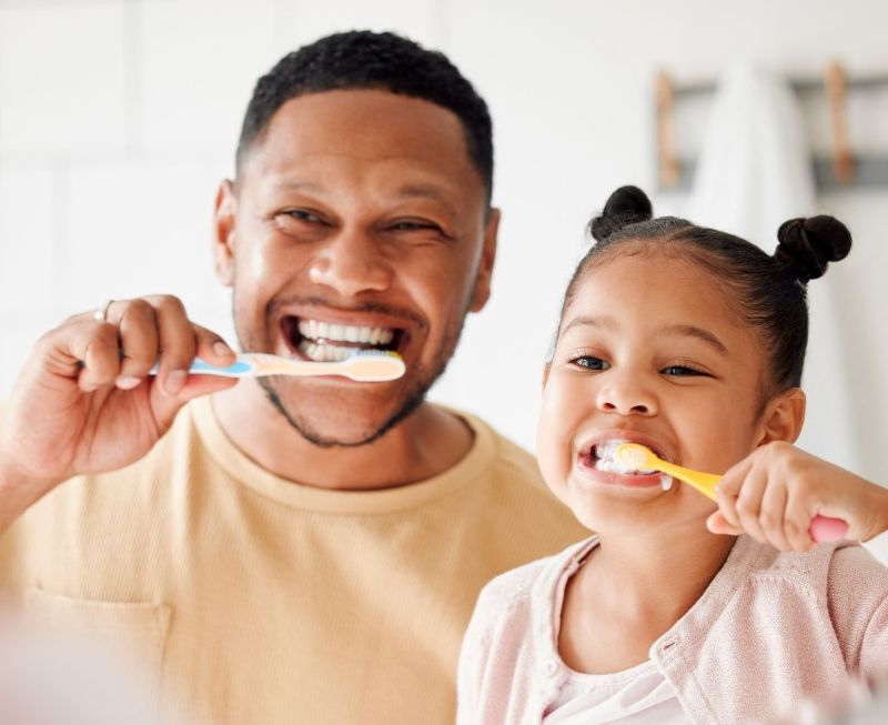 A man and a little girl are brushing their teeth in front of a mirror.
