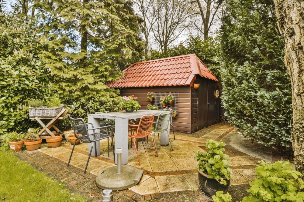 Patio with table, chairs, garden shed, and surrounding greenery. Red tiled roof on shed.