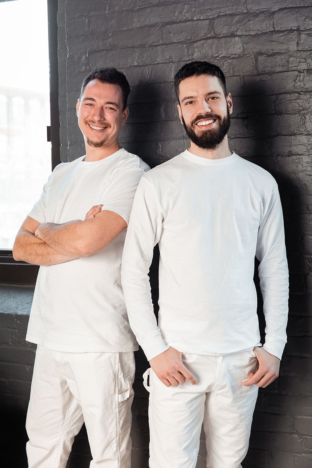 Deux personnes souriantes vêtues de blanc se tiennent devant un mur de briques sombres.