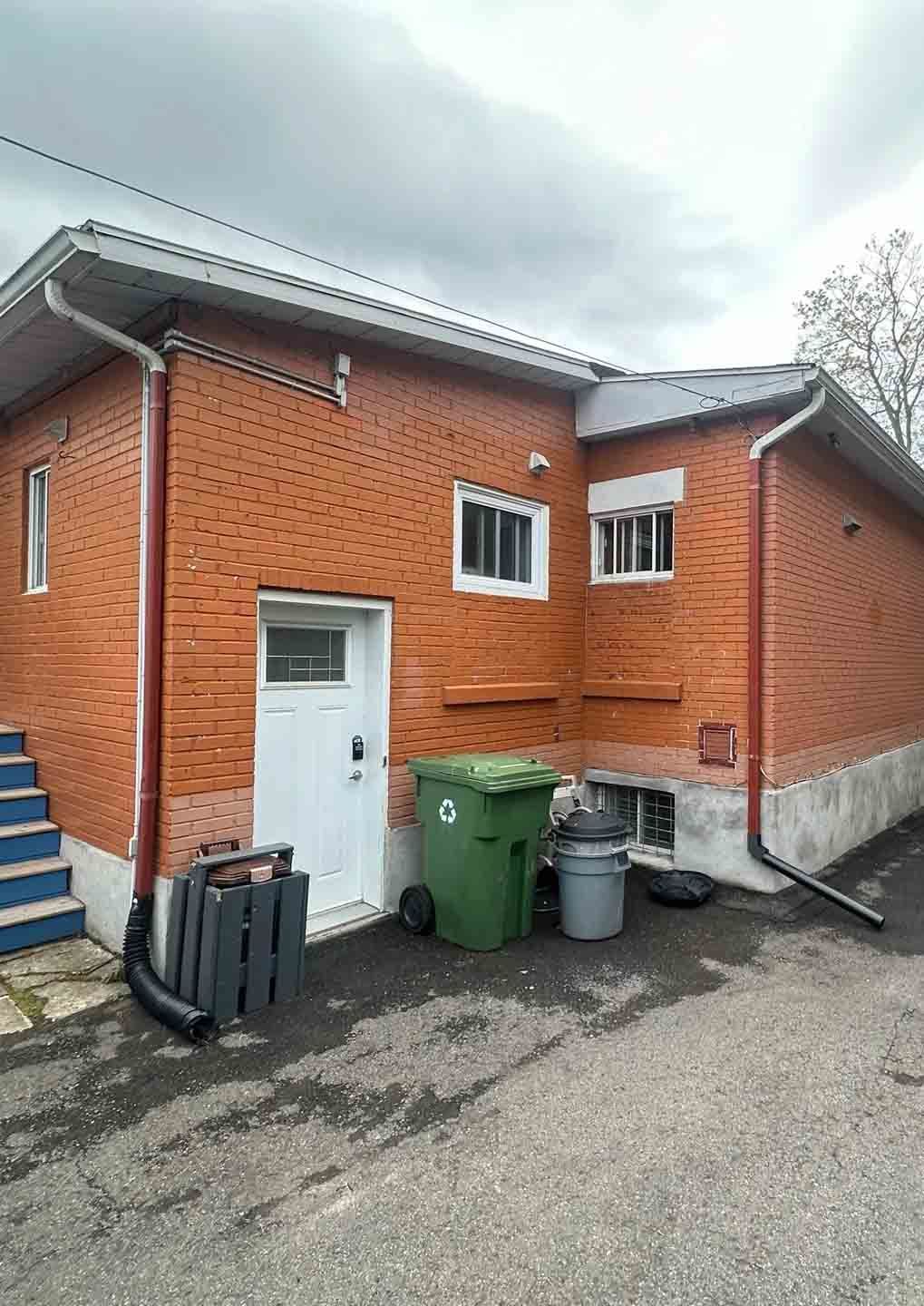 Bâtiment en briques avec porte et fenêtres blanches, poubelles vertes et escalier bleu à côté d'une zone pavée.