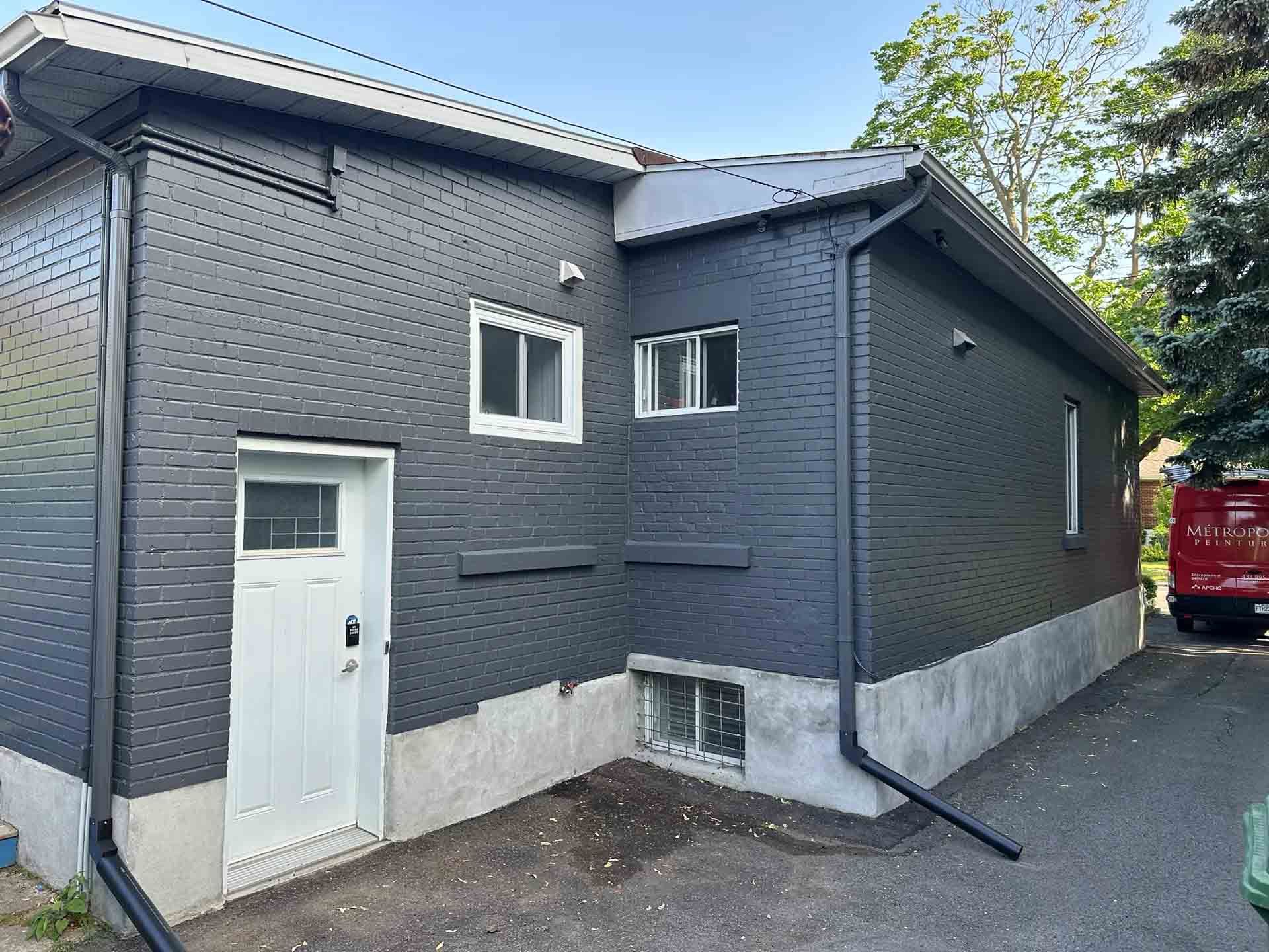 Maison en briques gris foncé avec porte, fenêtres et ventilation de sous-sol blanches, située à côté d'une allée et d'un arbre.
