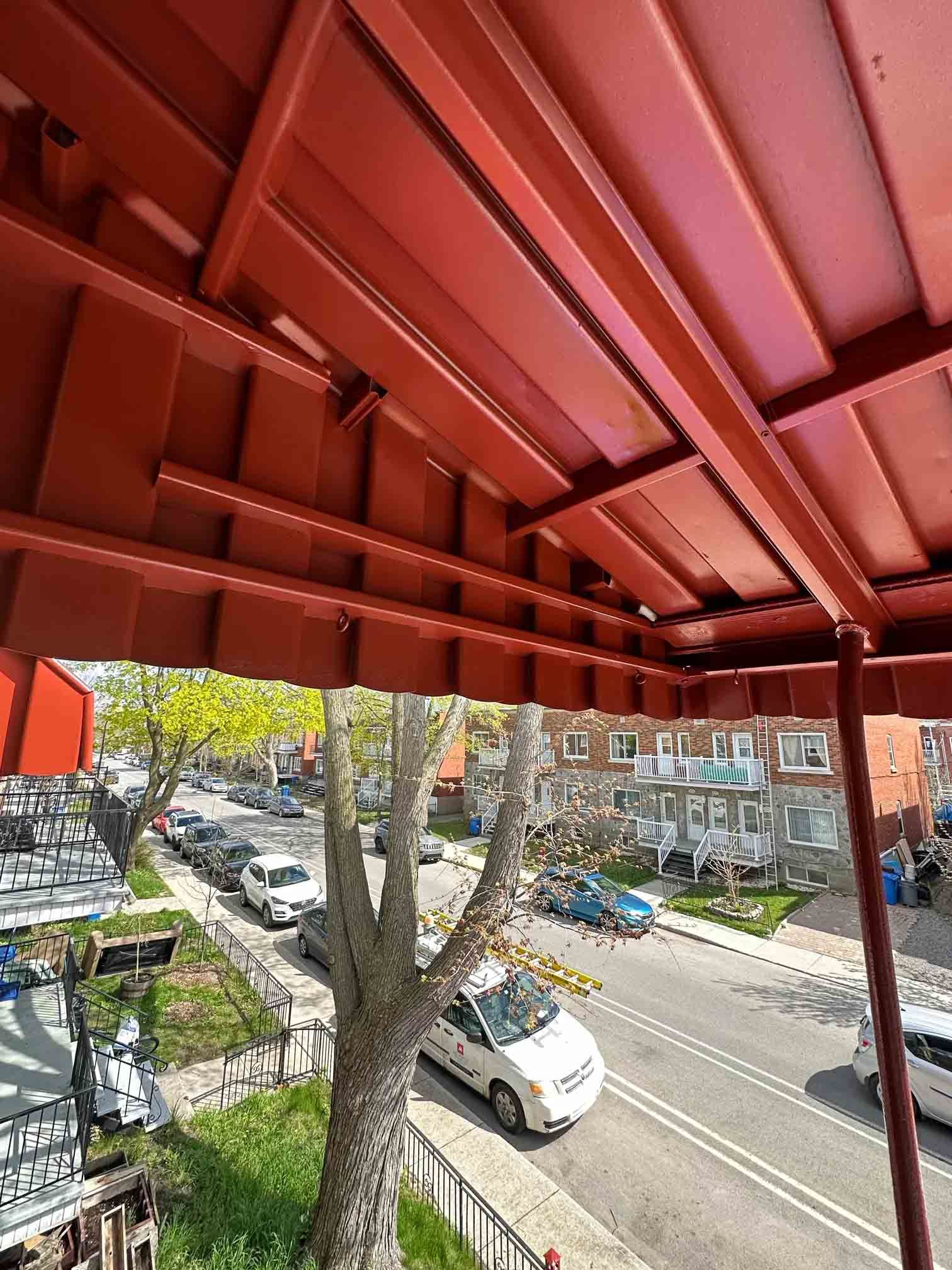 Auvent rouge donnant sur une rue bordée d'arbres, avec des voitures garées et des immeubles d'appartements en contrebas.