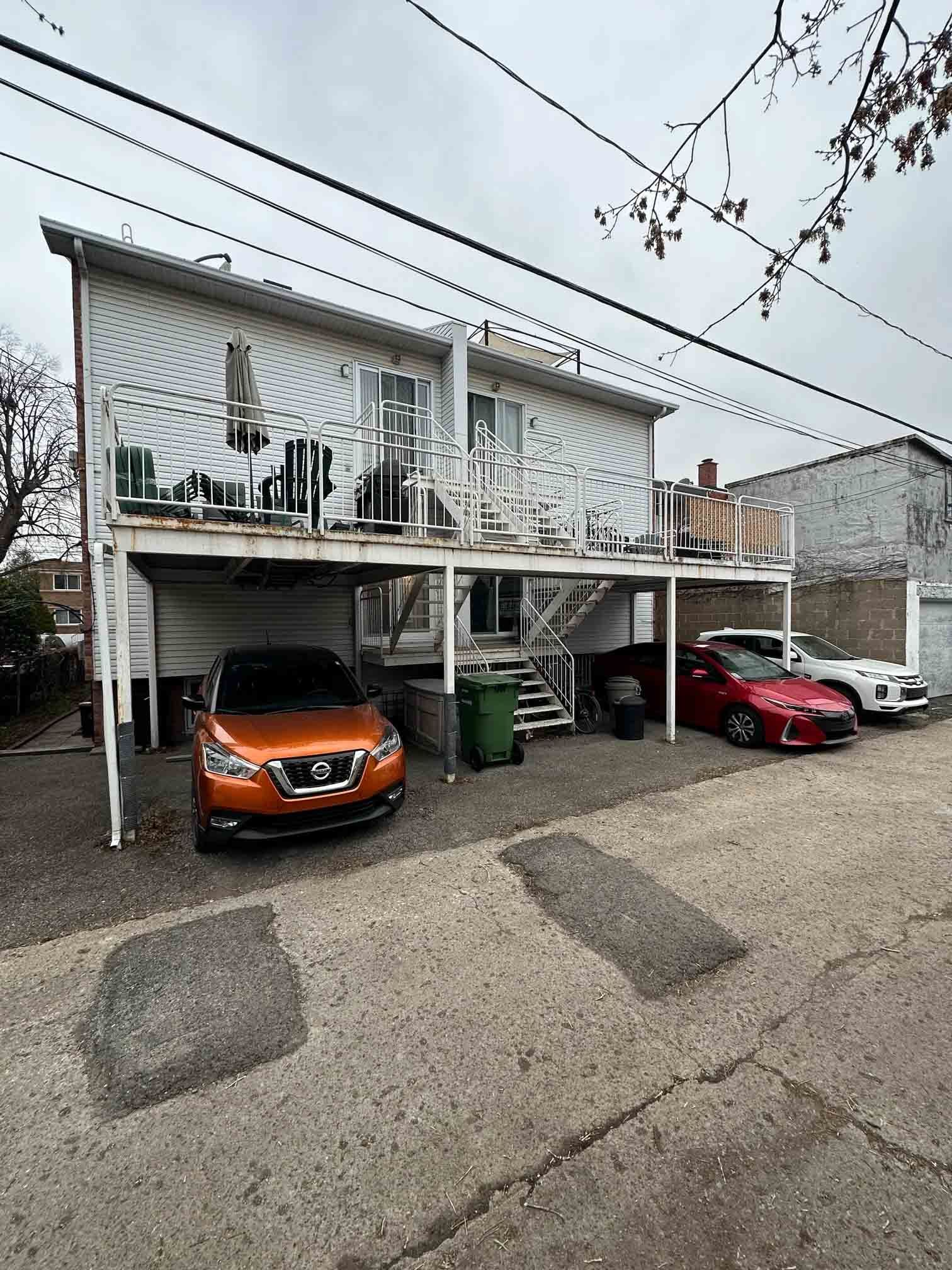 Duplex à deux étages avec des voitures garées en dessous, dont un SUV orange et une berline rouge, par temps nuageux