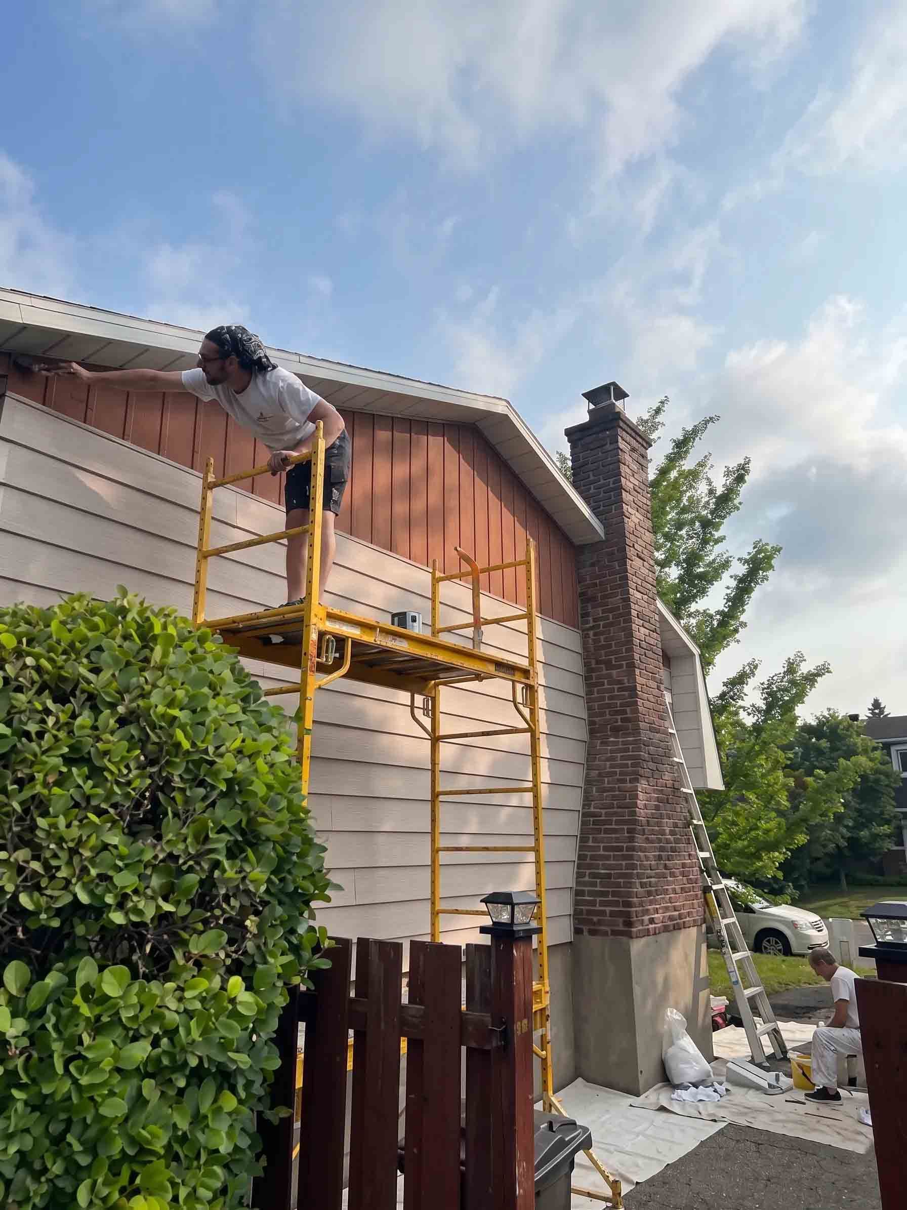 Ouvrier sur un échafaudage en train de peindre l'extérieur d'une maison à côté d'une cheminée en briques sous un ciel bleu