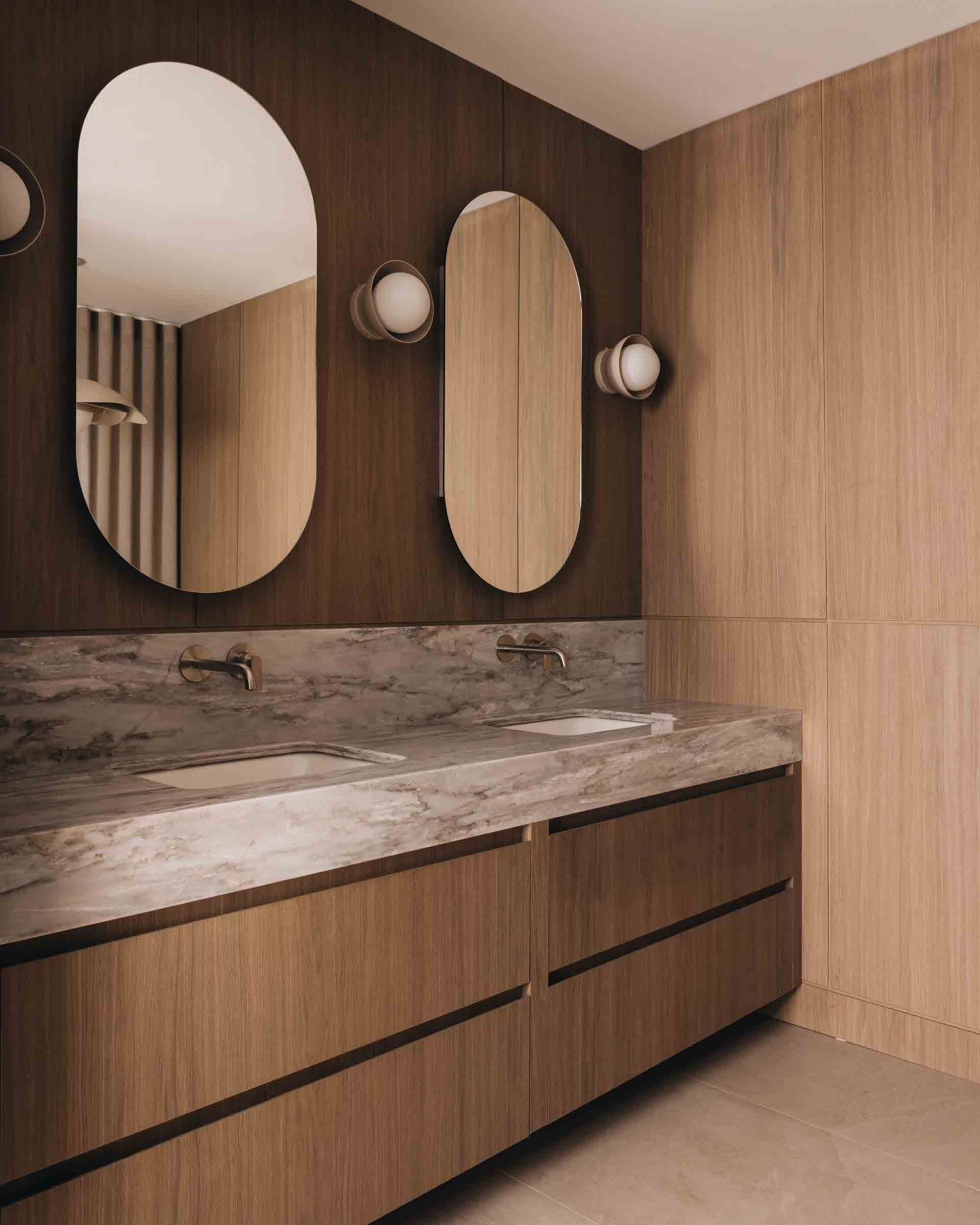 Meuble-lavabo moderne avec comptoir en marbre, armoires en bois et trois miroirs ovales sur un mur lambrissé.
