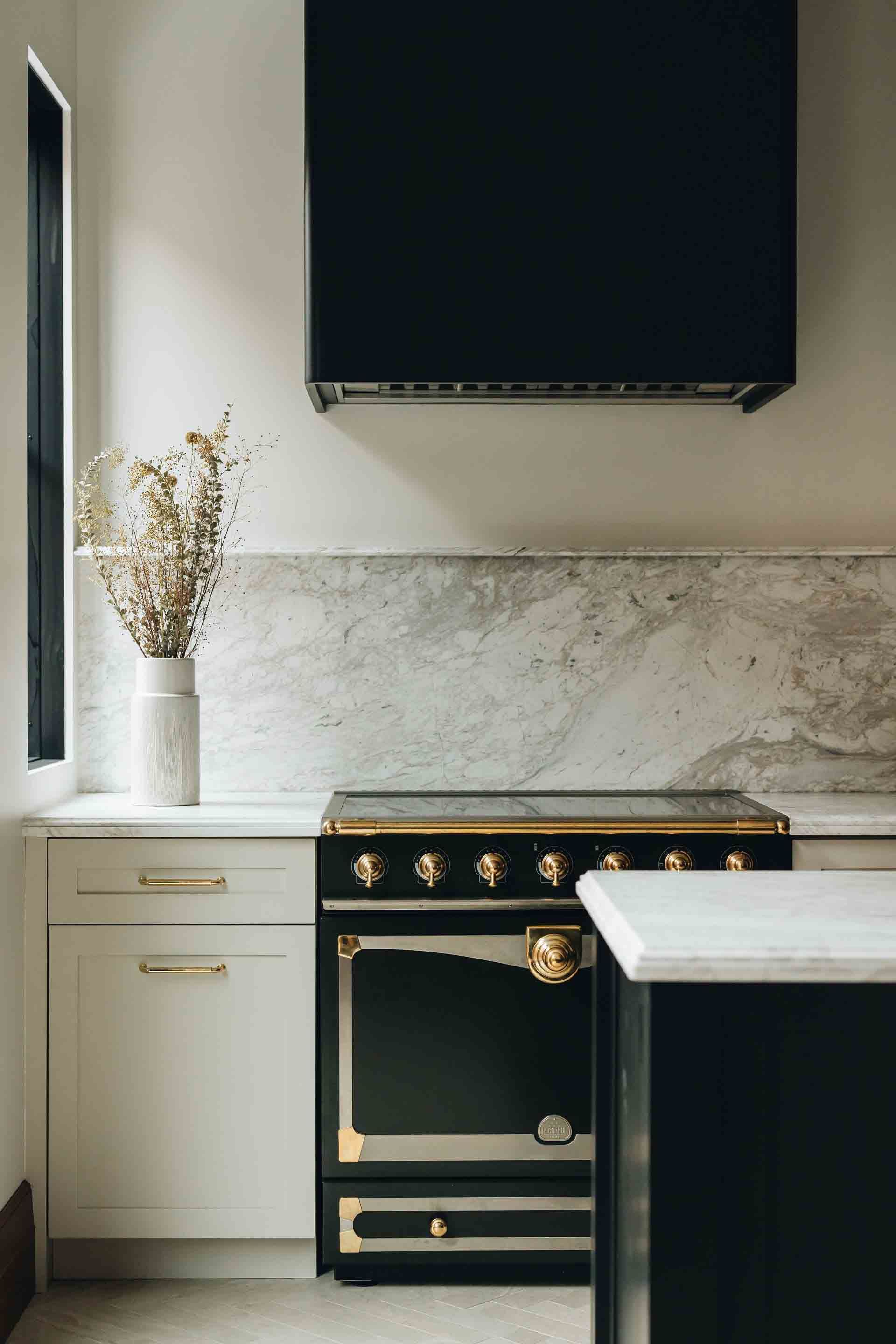 Cuisine moderne avec cuisinière noire, placards blancs, crédence en marbre et vase de fleurs séchées près de la fenêtre.