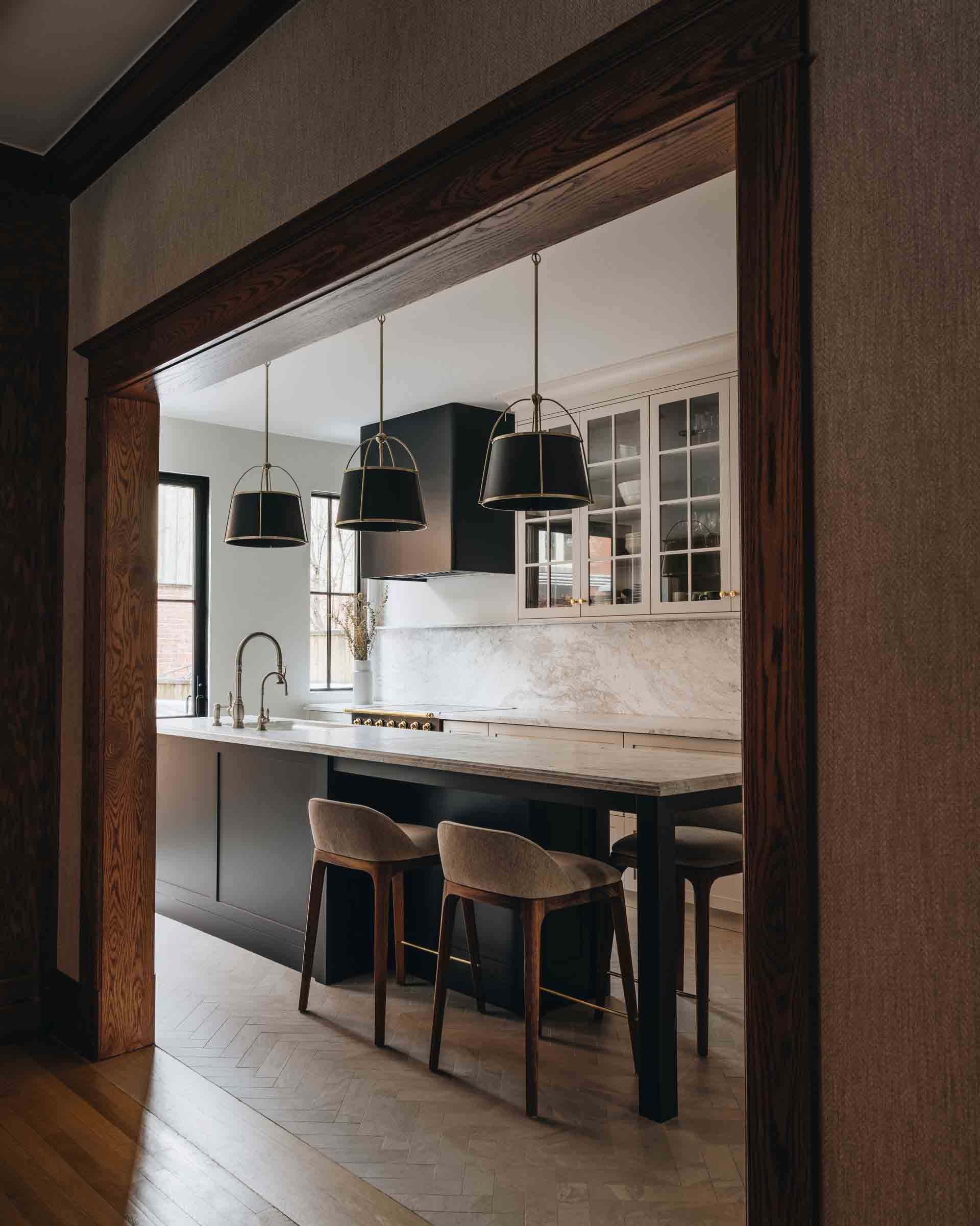 Cuisine moderne vue à travers une porte, avec îlot central foncé, suspensions lumineuses et tabourets de bar sur parquet.