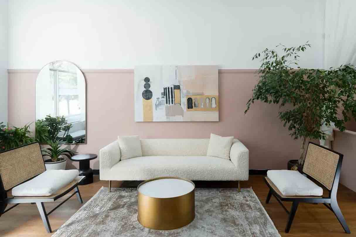 Salon lumineux avec canapé blanc, table basse dorée, murs roses, plantes et art mural abstrait.