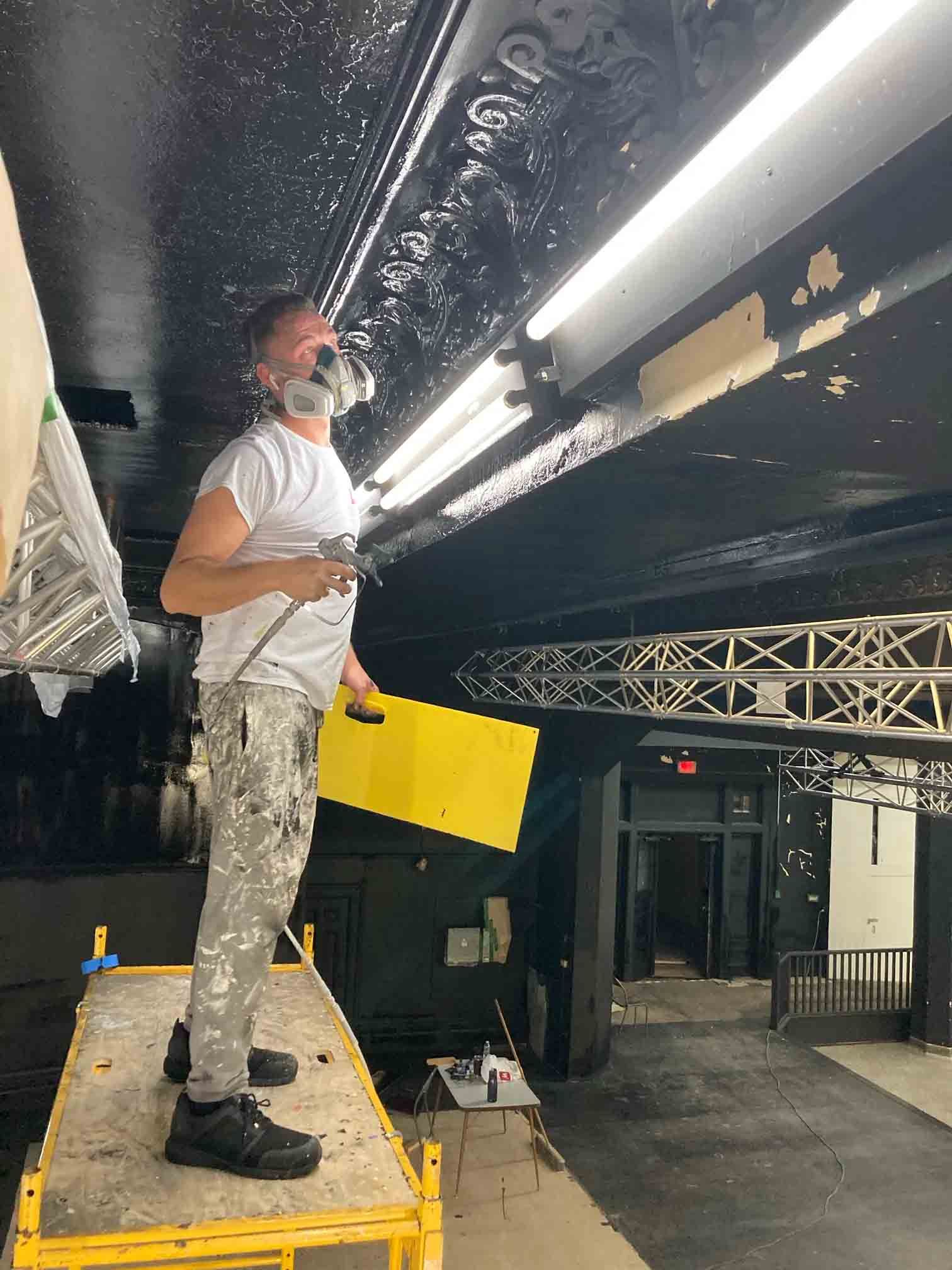 Une personne se trouve sur une plateforme jaune dans un espace intérieur sombre, travaillant près de luminaires au plafond et de structures en treillis.