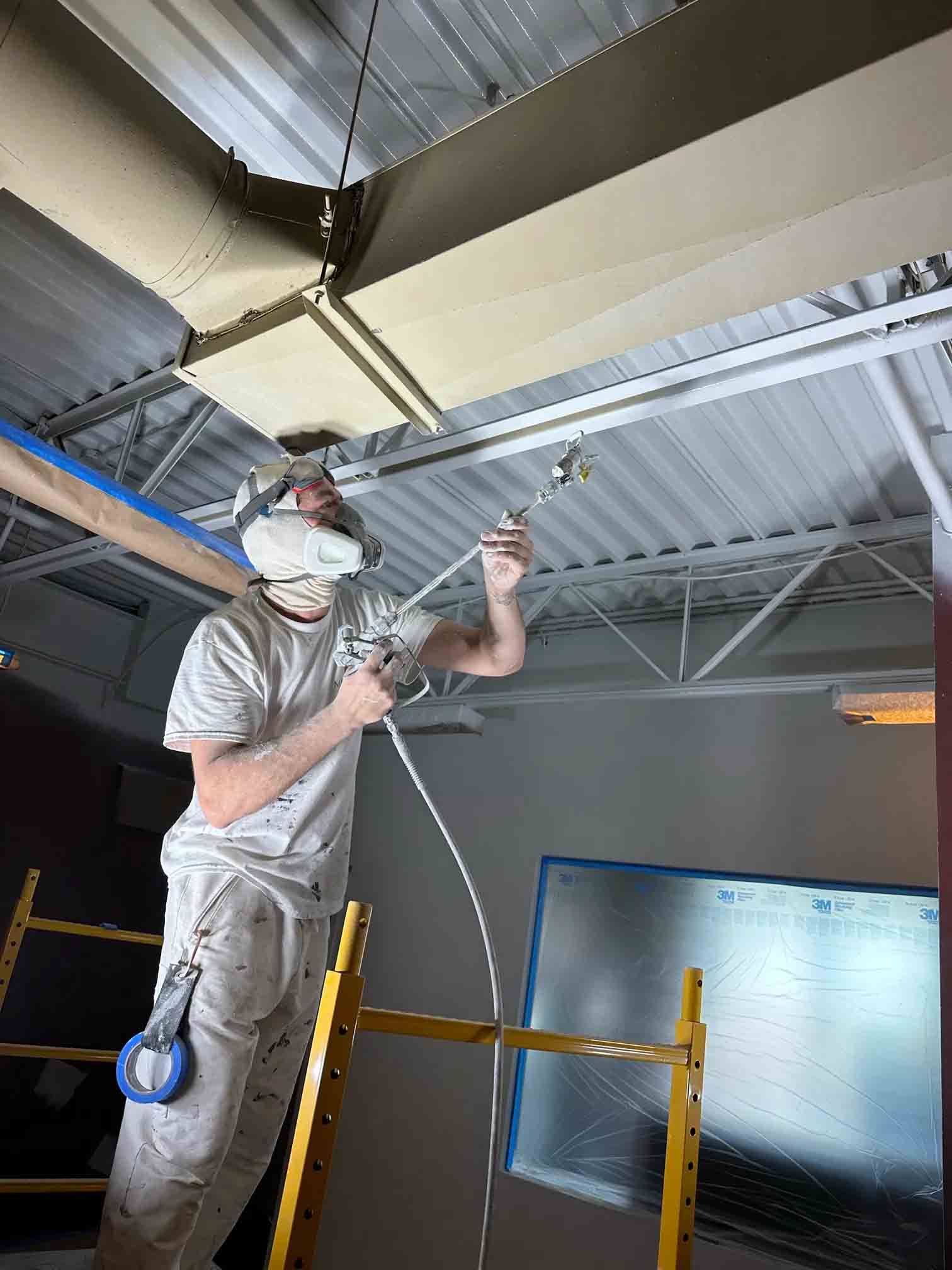 Un ouvrier portant un masque peint à la bombe une poutre de plafond à l'intérieur d'une pièce encombrée d'échafaudages.