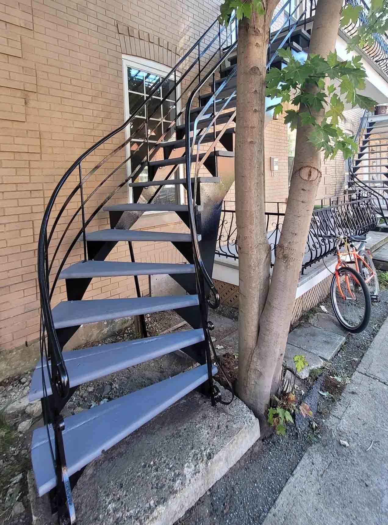 Escalier en colimaçon en métal noir à côté d'un bâtiment en briques, avec un arbre et un vélo à proximité.