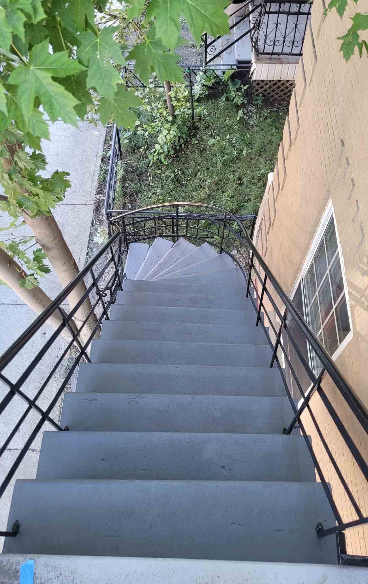 Escalier extérieur abrupt avec rampes noires, longeant un mur de briques et des arbres feuillus.