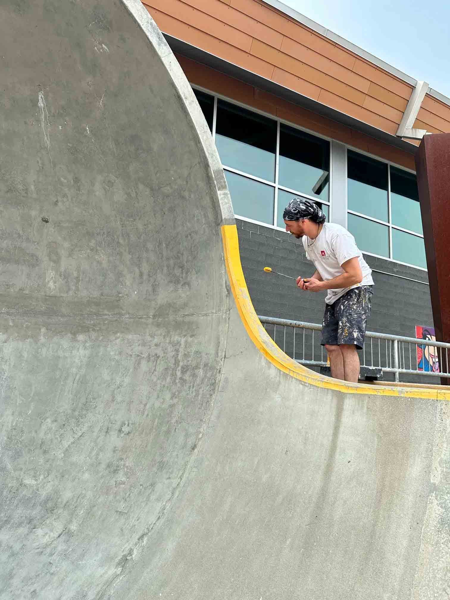 Une personne ponce un bowl de skate en béton avec un outil portatif à l'extérieur, près d'un bâtiment.