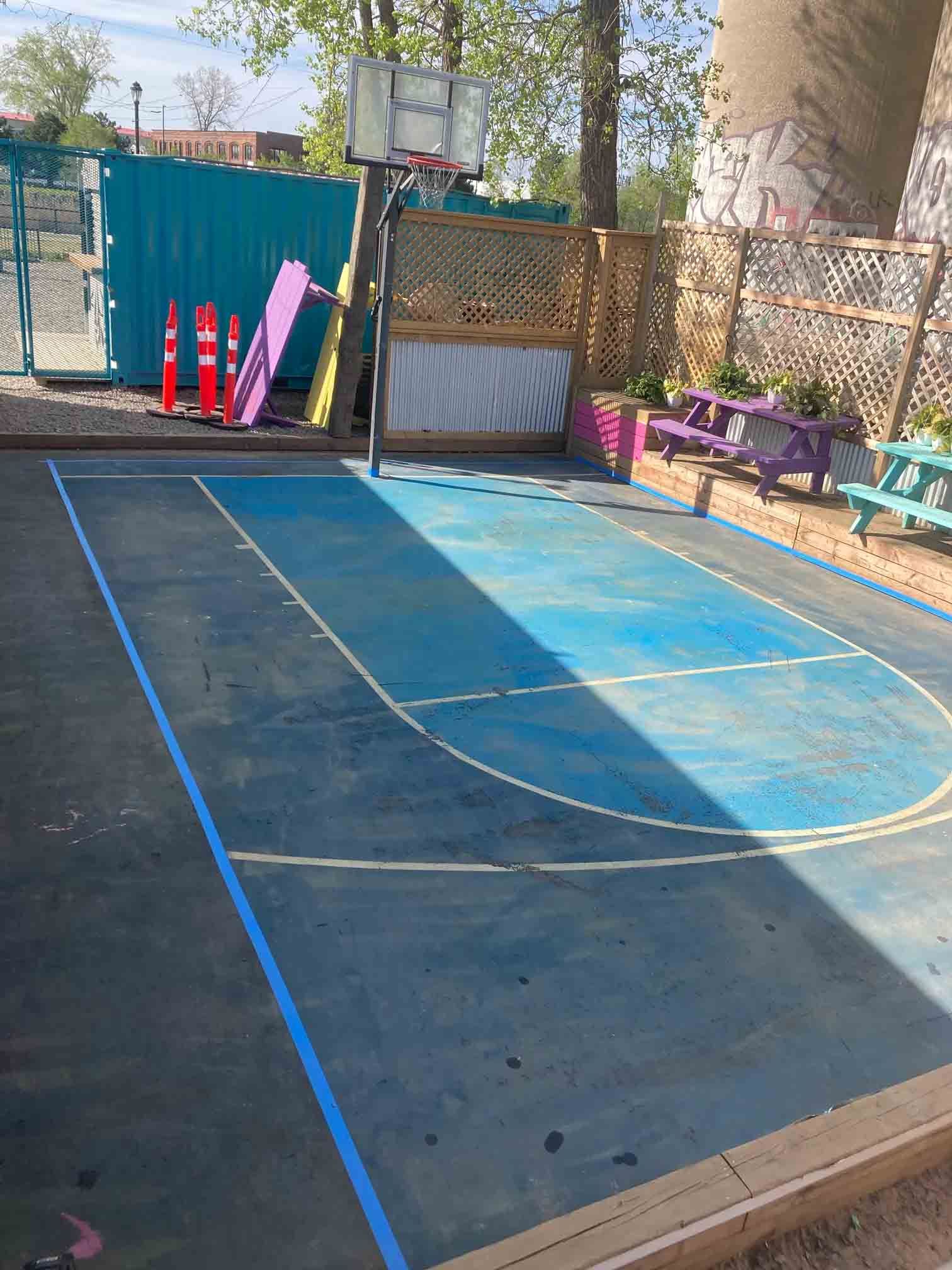 Terrain extérieur avec revêtement bleu, cercle jaune et équipements de jeux le long d'une clôture à l'ombre tachetée