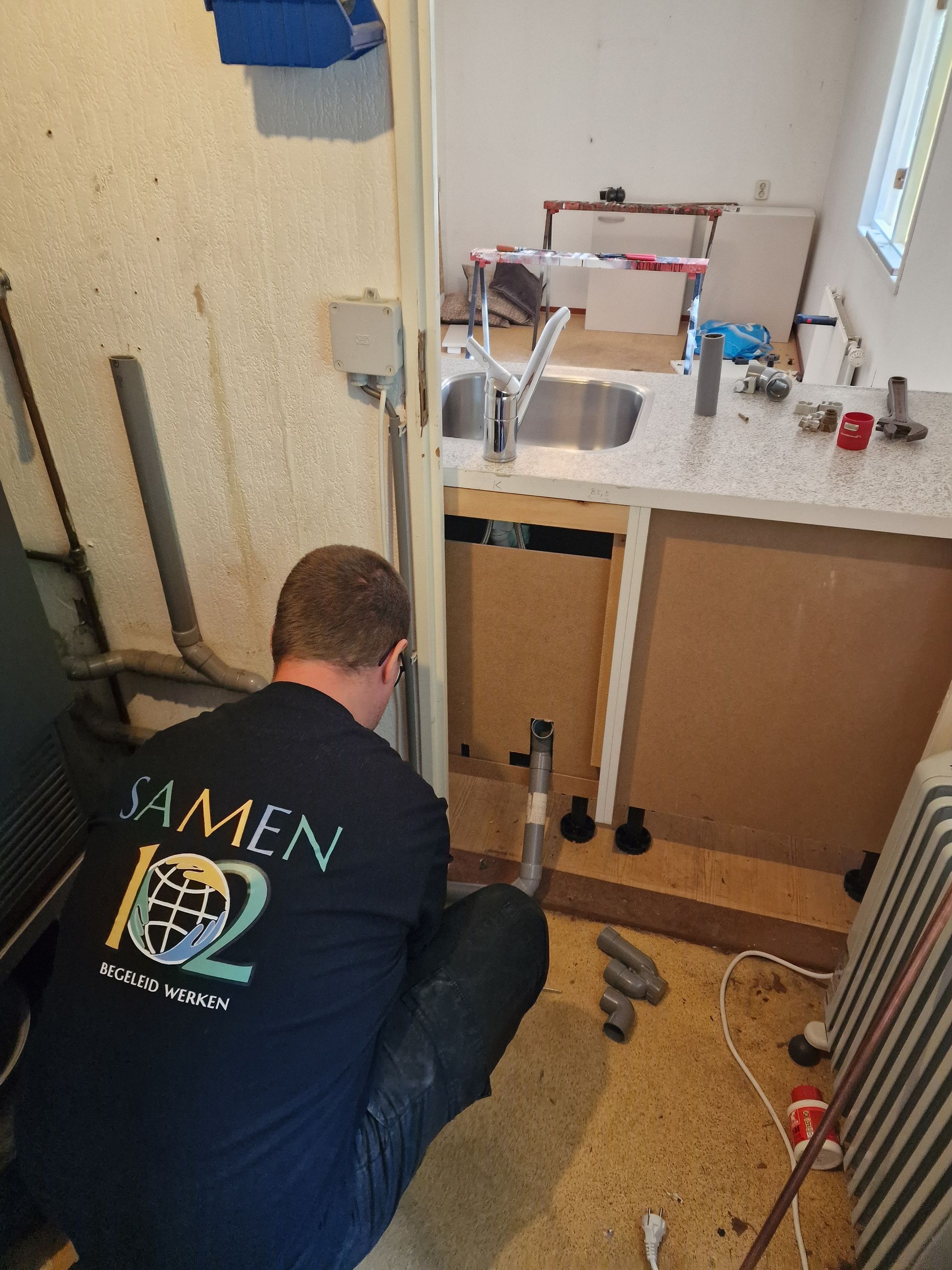 Een man knielt in de keuken en werkt aan de gootsteen. Dagbesteding Samen102 Putten