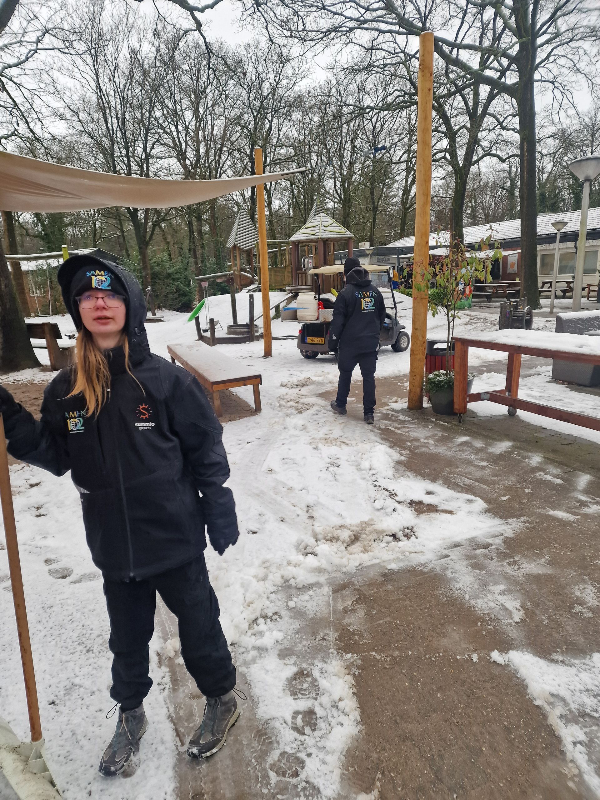 Persoon in zwarte jas en broek met een stok in de hand, staand op besneeuwde grond; een andere persoon in de verte; buitenomgeving.