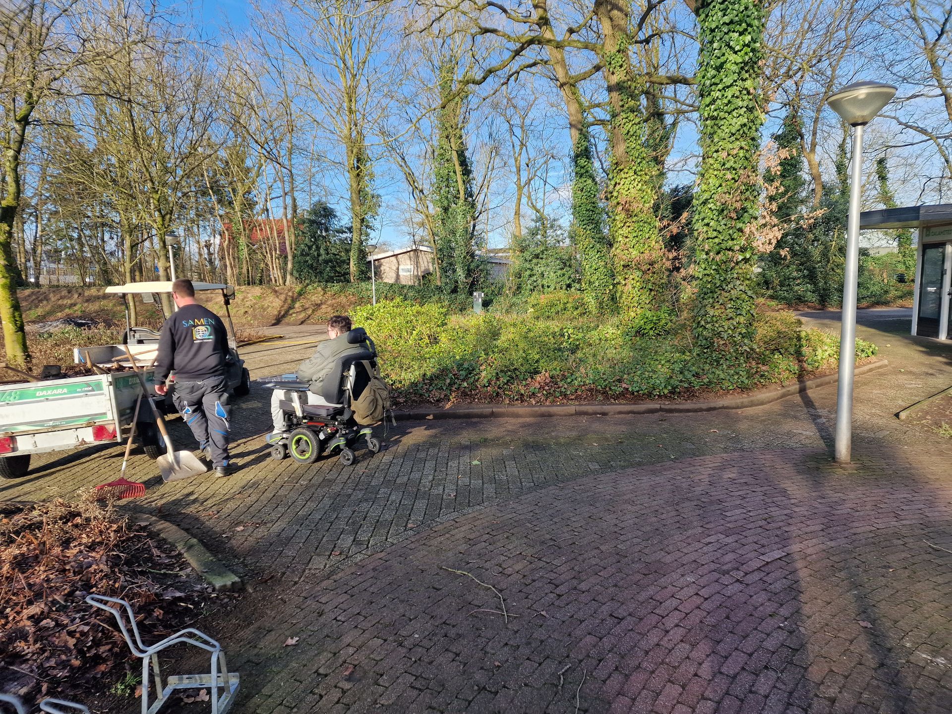 Twee arbeiders maken een met bakstenen bestraat gebied schoon in de buurt van een trailer en wat begroeiing. Zonnige dag, kale bomen.