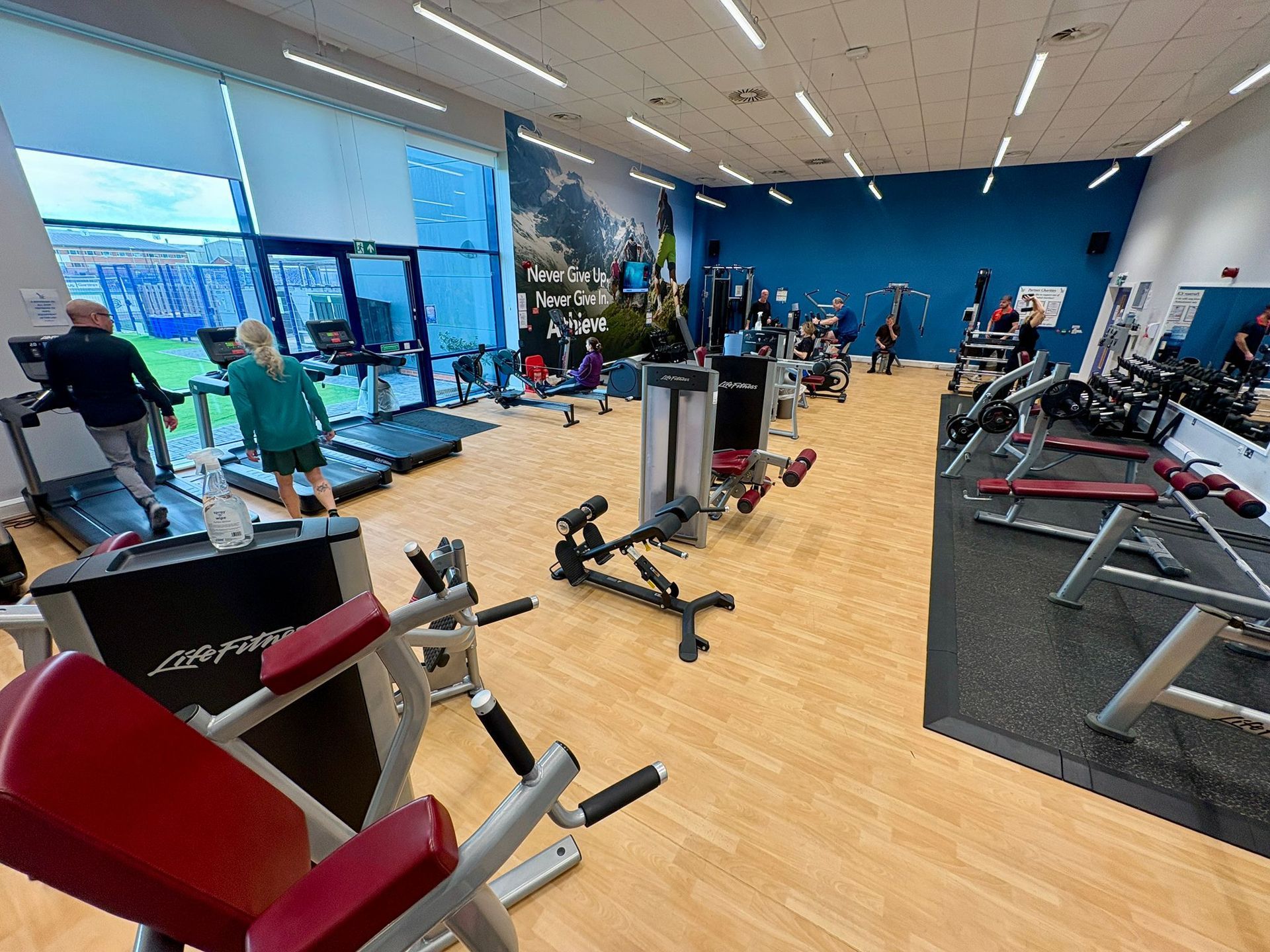 Gym interior with workout machines, people exercising near a large window, blue walls.