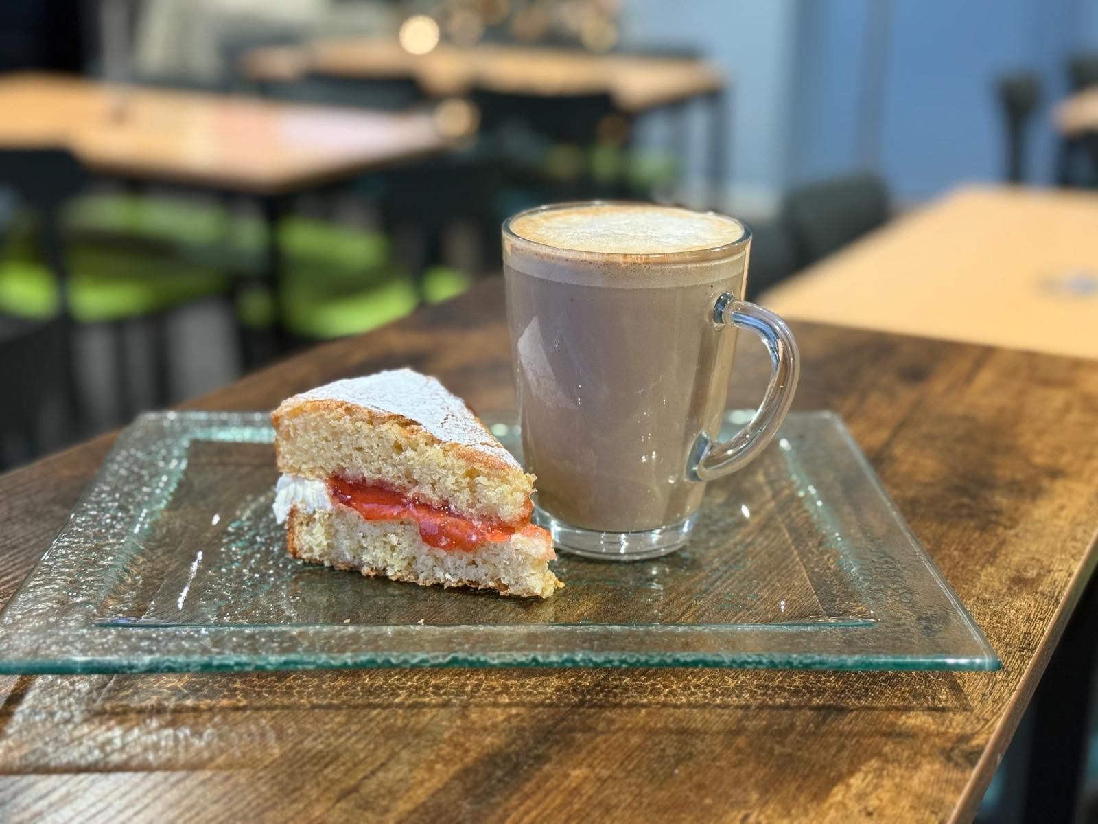 Slice of cake with a cream and jam filling next to a coffee drink, on a glass plate, on a wooden table.