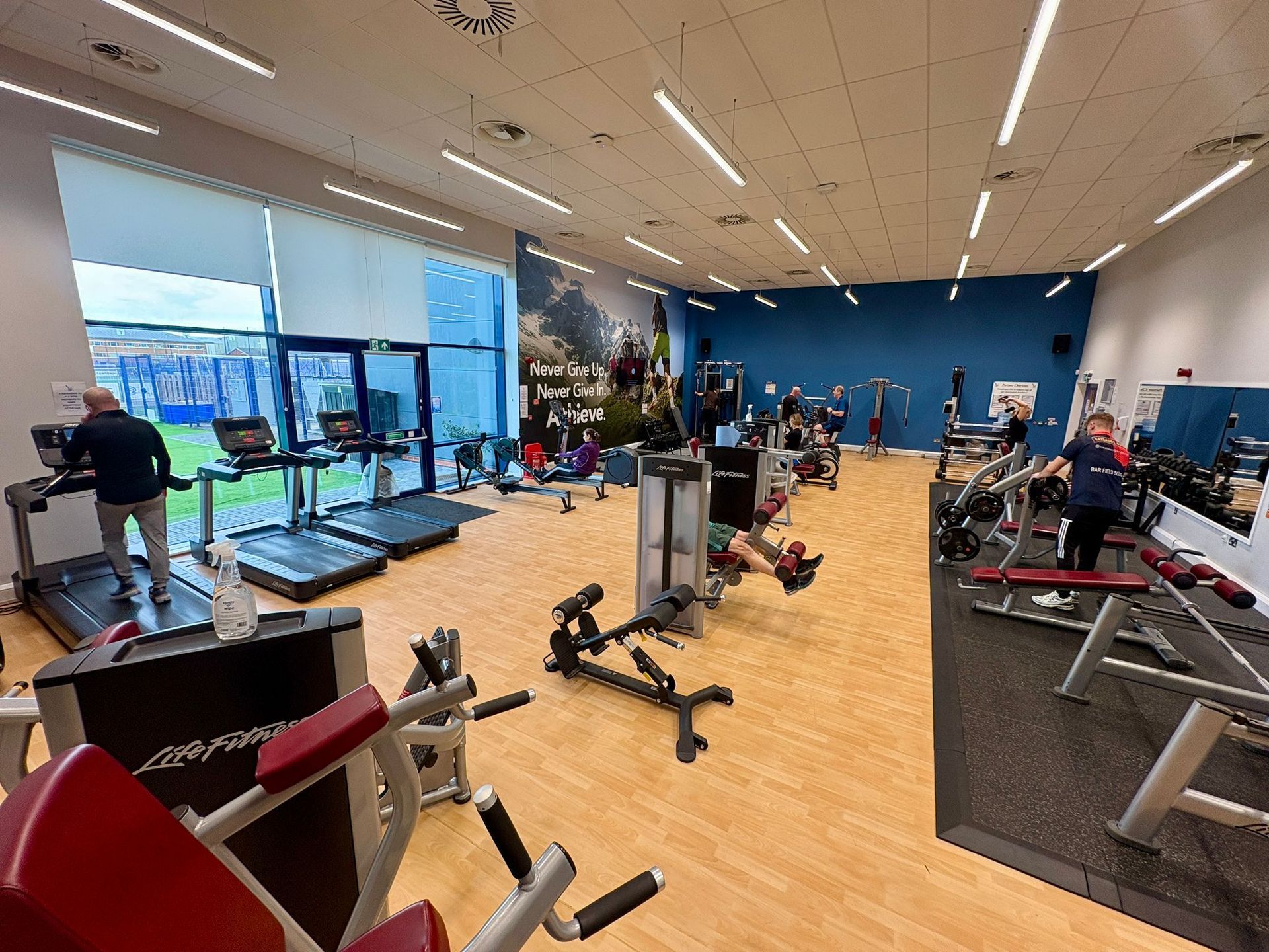 Gym interior with treadmills, weight machines, and people exercising; blue and white walls, wooden floor.