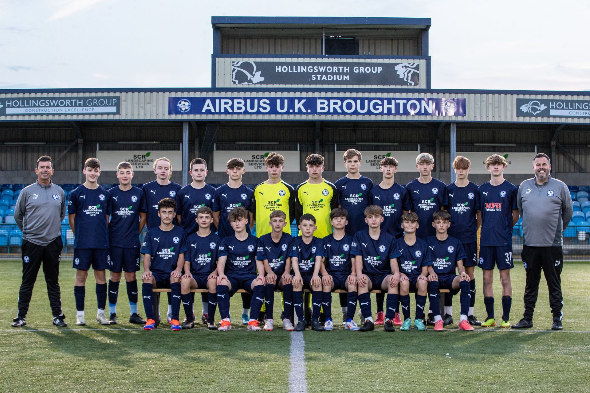 Airbus UK Broughton soccer team posing on a field in blue uniforms. Stadium background.