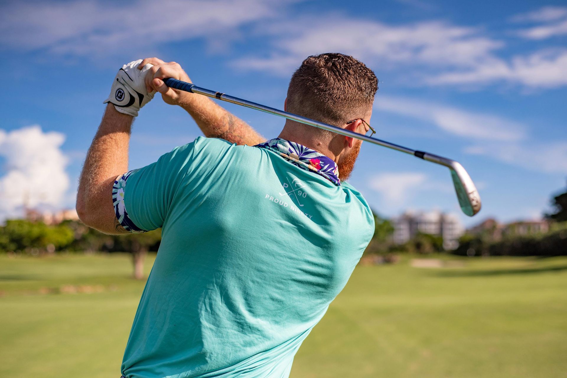 Golfer in turquoise shirt swings club on green course under a blue sky.