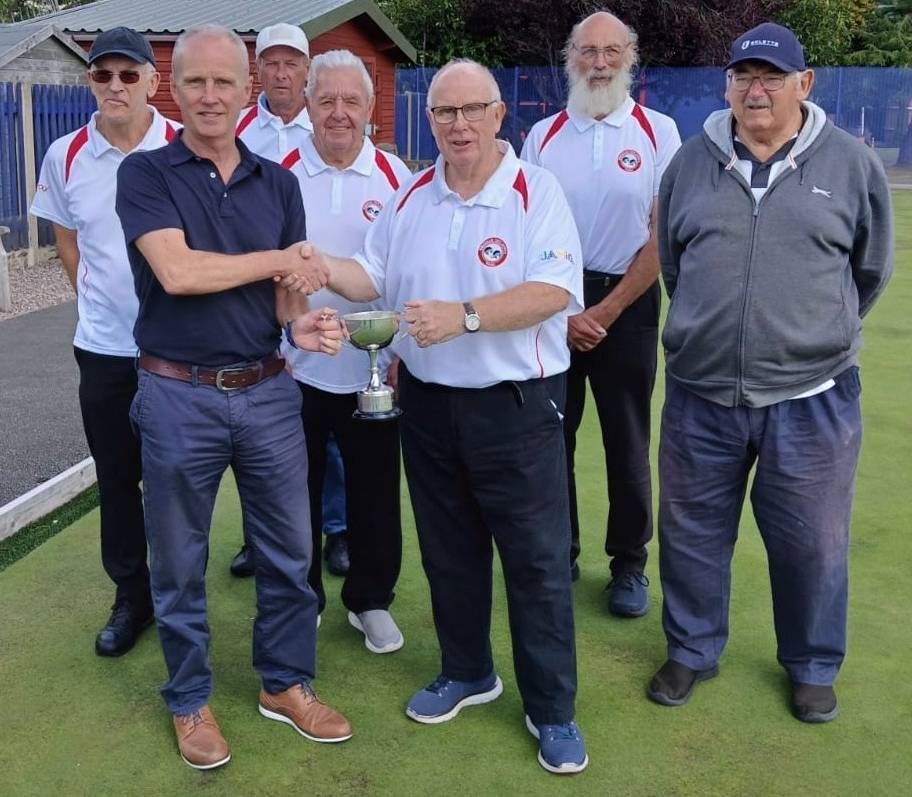 Group of men on a lawn; one shaking hands, another holding a trophy, others in matching white shirts.