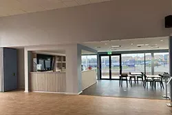 Interior view of a light-filled space with a counter area, seating, and a view of water through large windows.