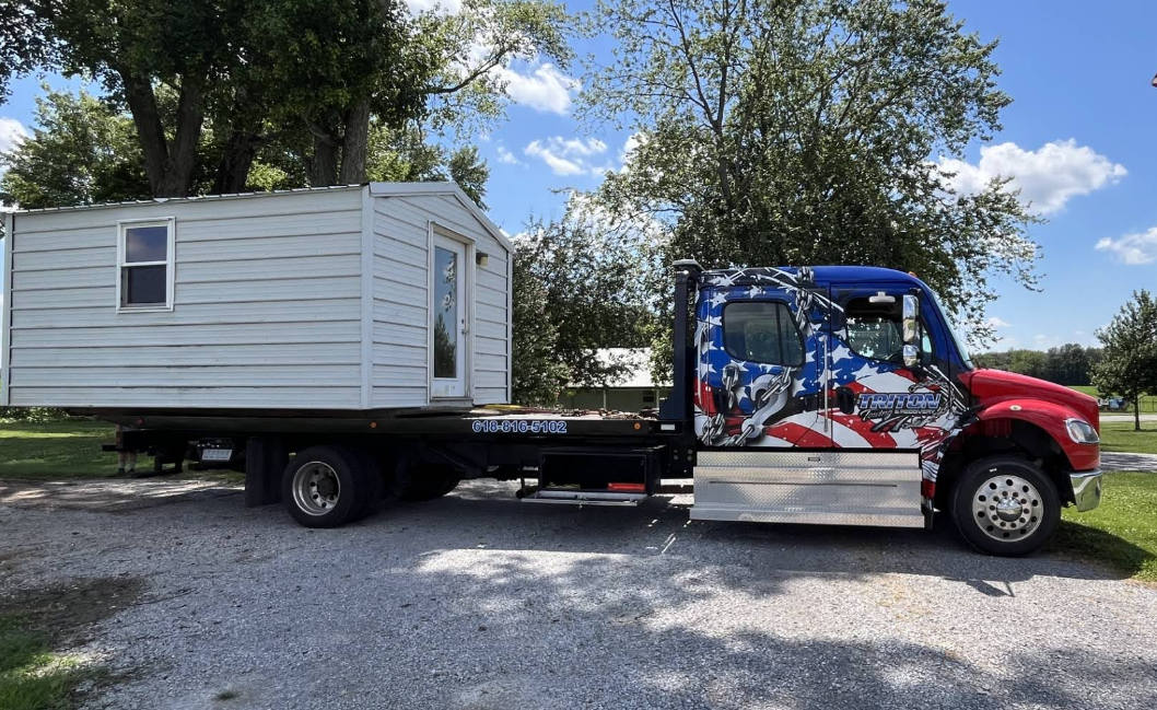 A small, white house is on a flatbed truck with an American flag paint job on the cab.