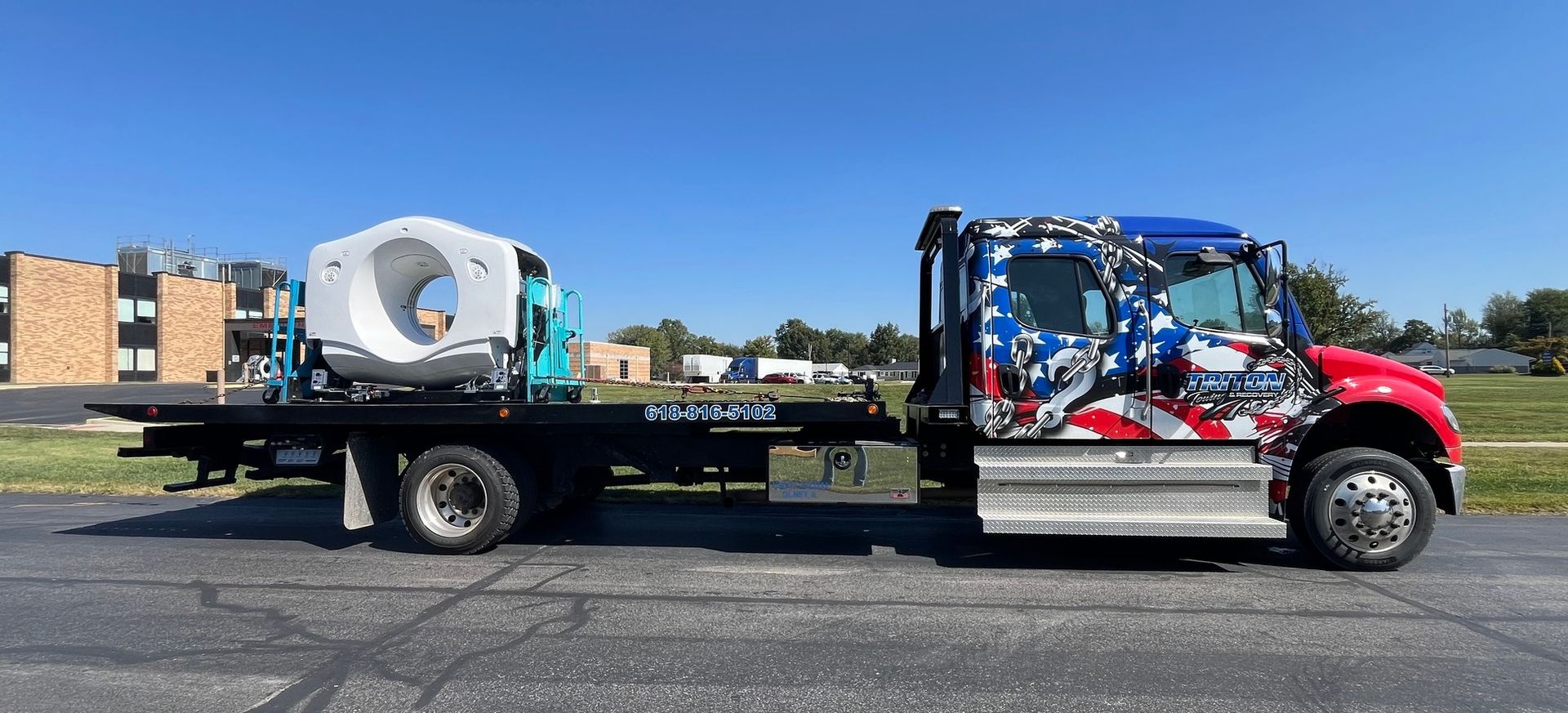 A flatbed tow truck with an American flag design is transporting a large, white machine on a sunny day.