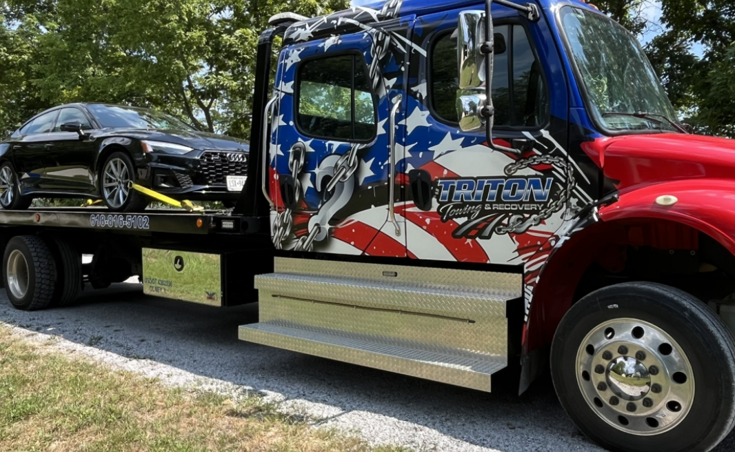 Tow truck with American flag graphics carrying a black car on a sunny day.