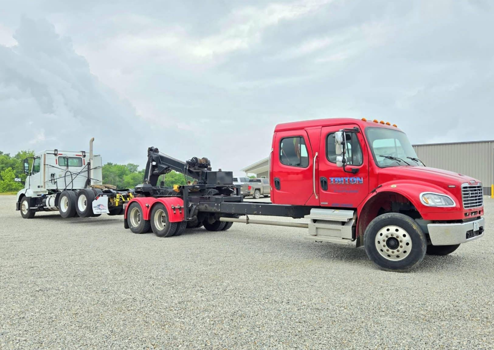 Red tow truck towing a white semi-truck on a gravel lot under a cloudy sky.