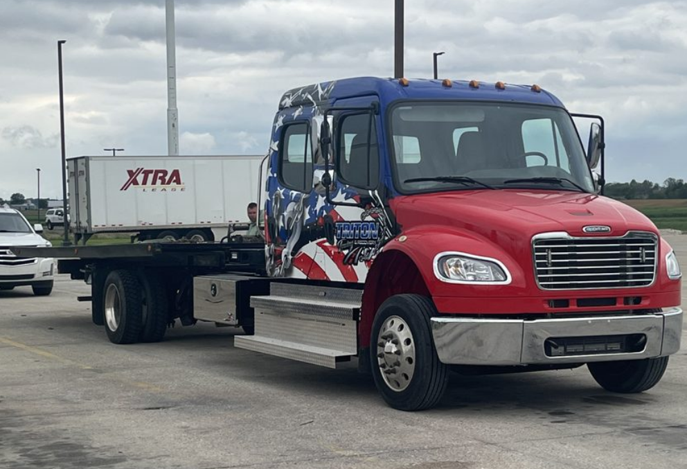 Red, white, and blue flatbed truck with patriotic graphics, carrying a white trailer labeled 