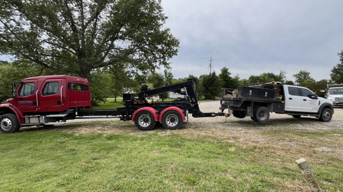 Red tow truck pulling a white flatbed truck on grass. Gray sky and trees in the background.