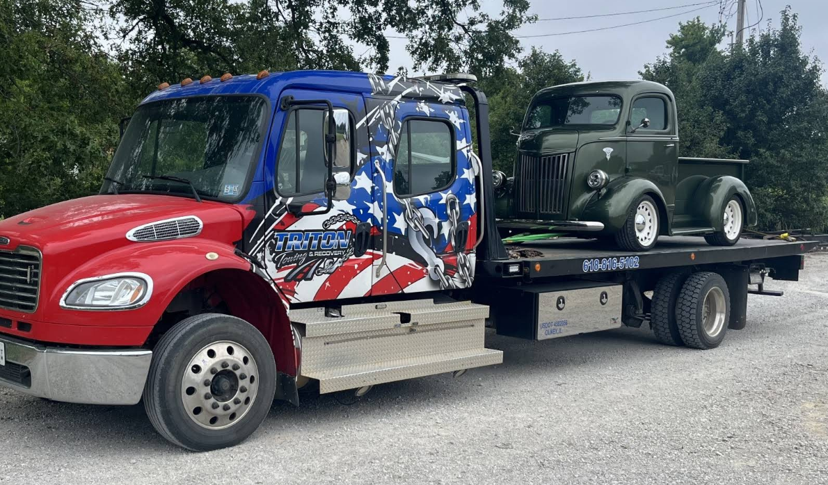 Red, white, and blue tow truck with a vintage green pickup truck on its flatbed. Outdoors.