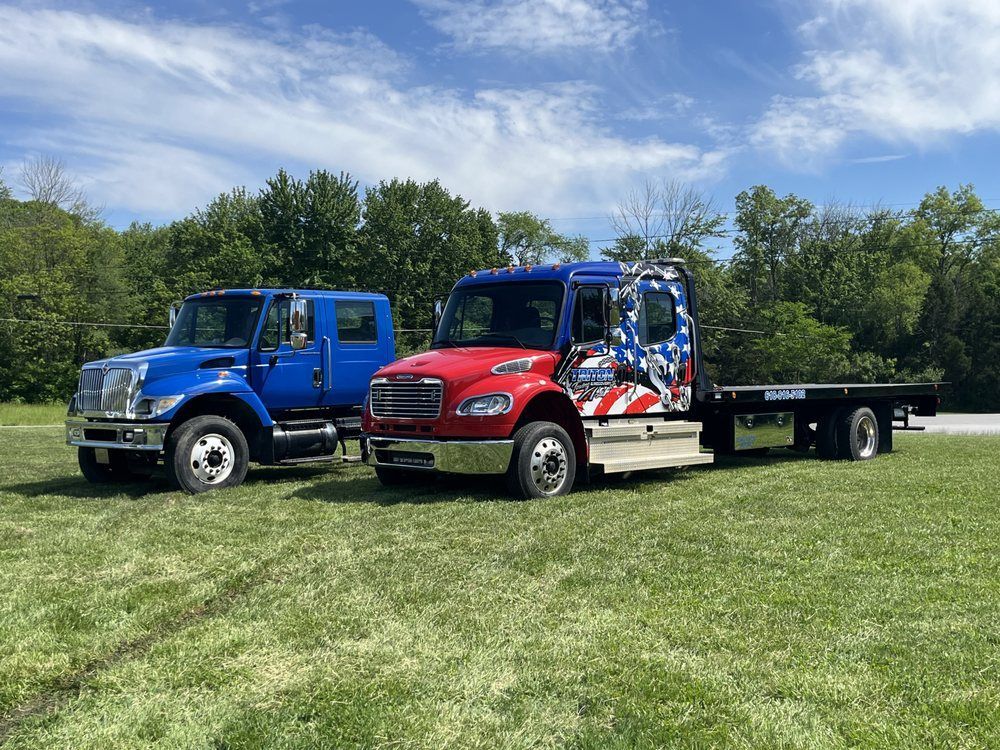 Two tow trucks, blue and red, parked on a grassy area with trees in the background. Sunny day.