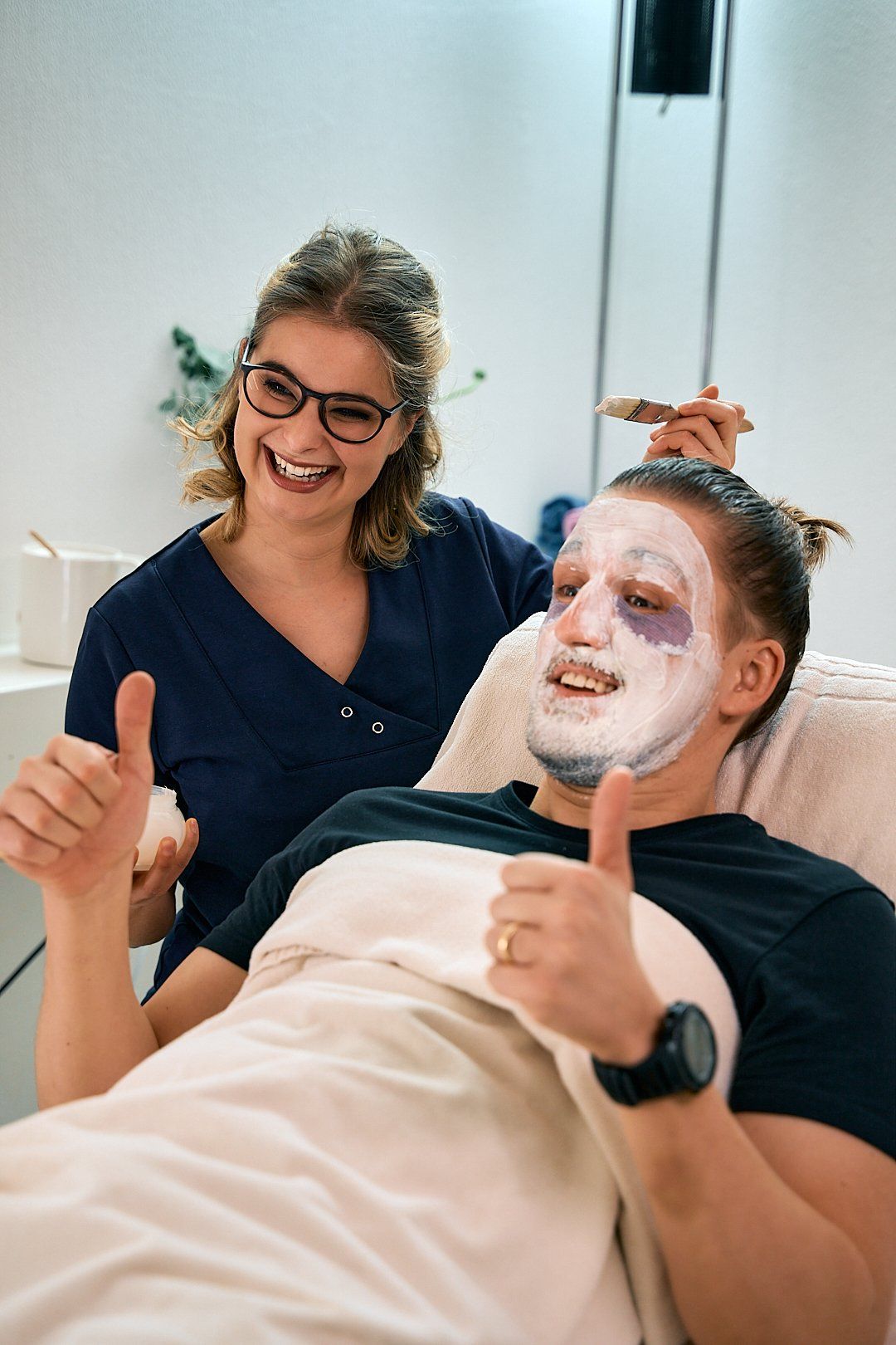Eine Kosmetikerin sitzt neben einem zufriedenen Kunden der eine Gesichtsmaske auf der Haut hat und die Daumen hoch hält