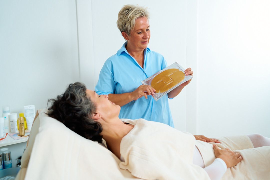 Kosmetikerin zeigt einer Kundin die auf der Spaliege liegt eine goldige Gesichtsmaske