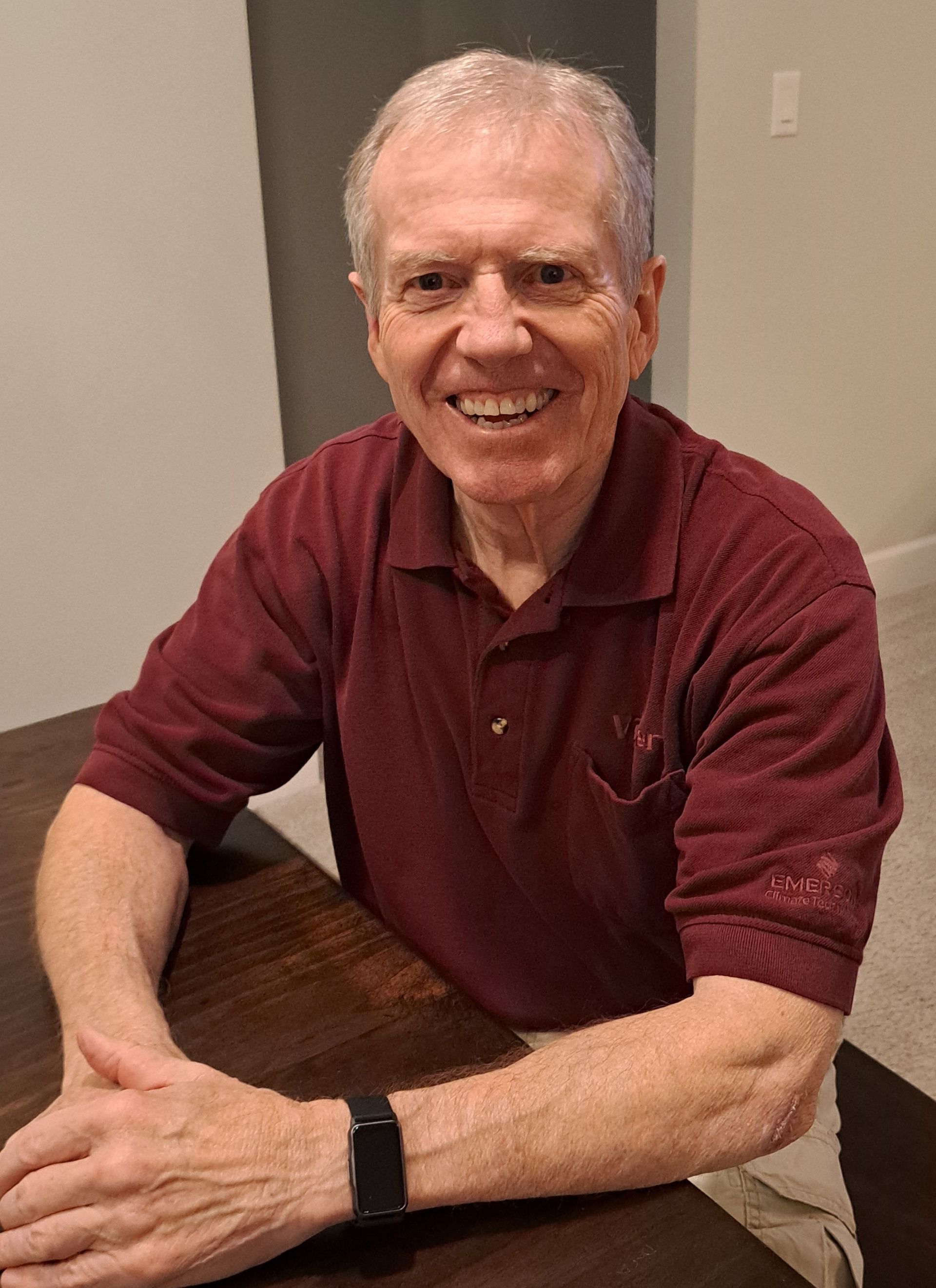 A man in a maroon shirt is sitting at a table with his hands folded and smiling.