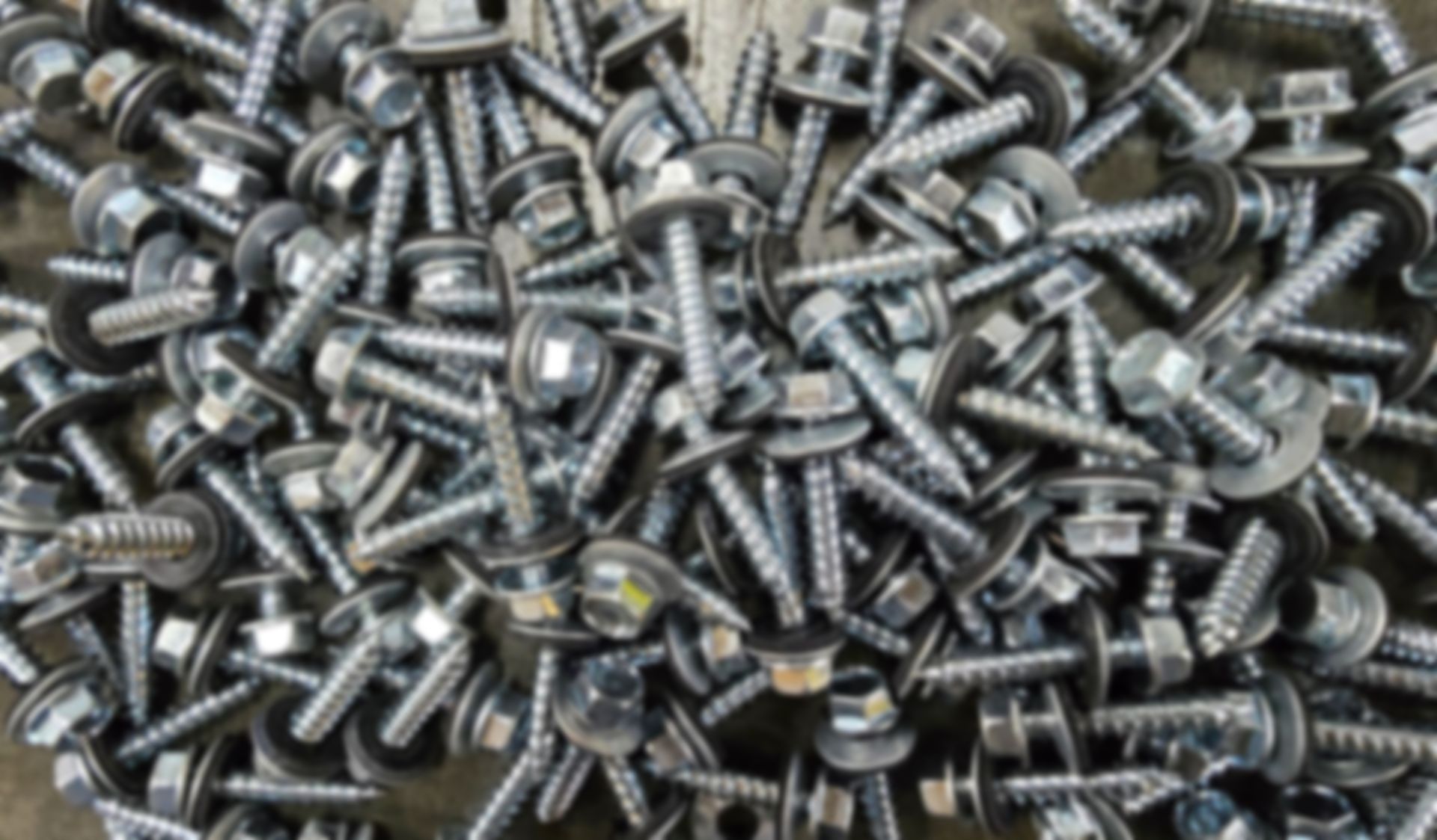 A pile of silver-colored metal roofing screws with hex washer heads and attached sealing washers.