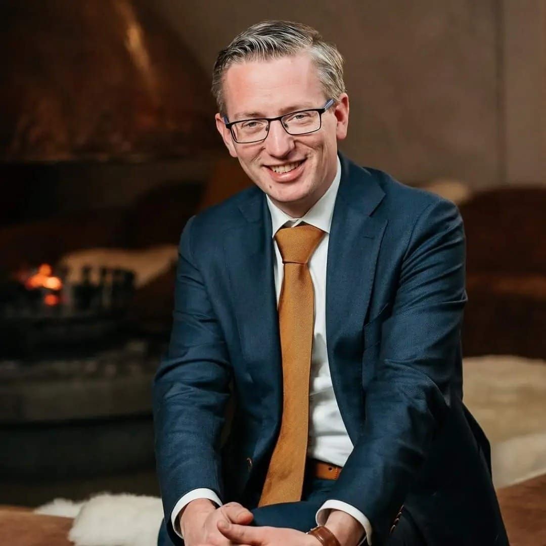 Man met bril en groene coltrui, lachend, voor een witte achtergrond.