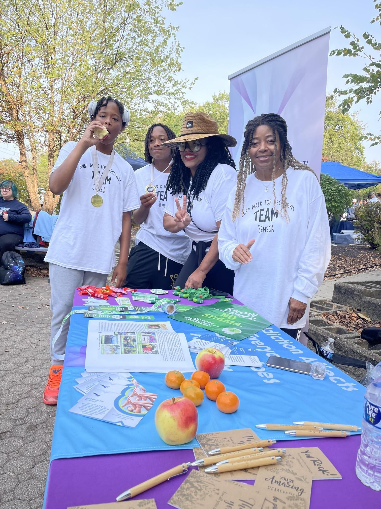 A group of people are standing around a table with apples and oranges on it.