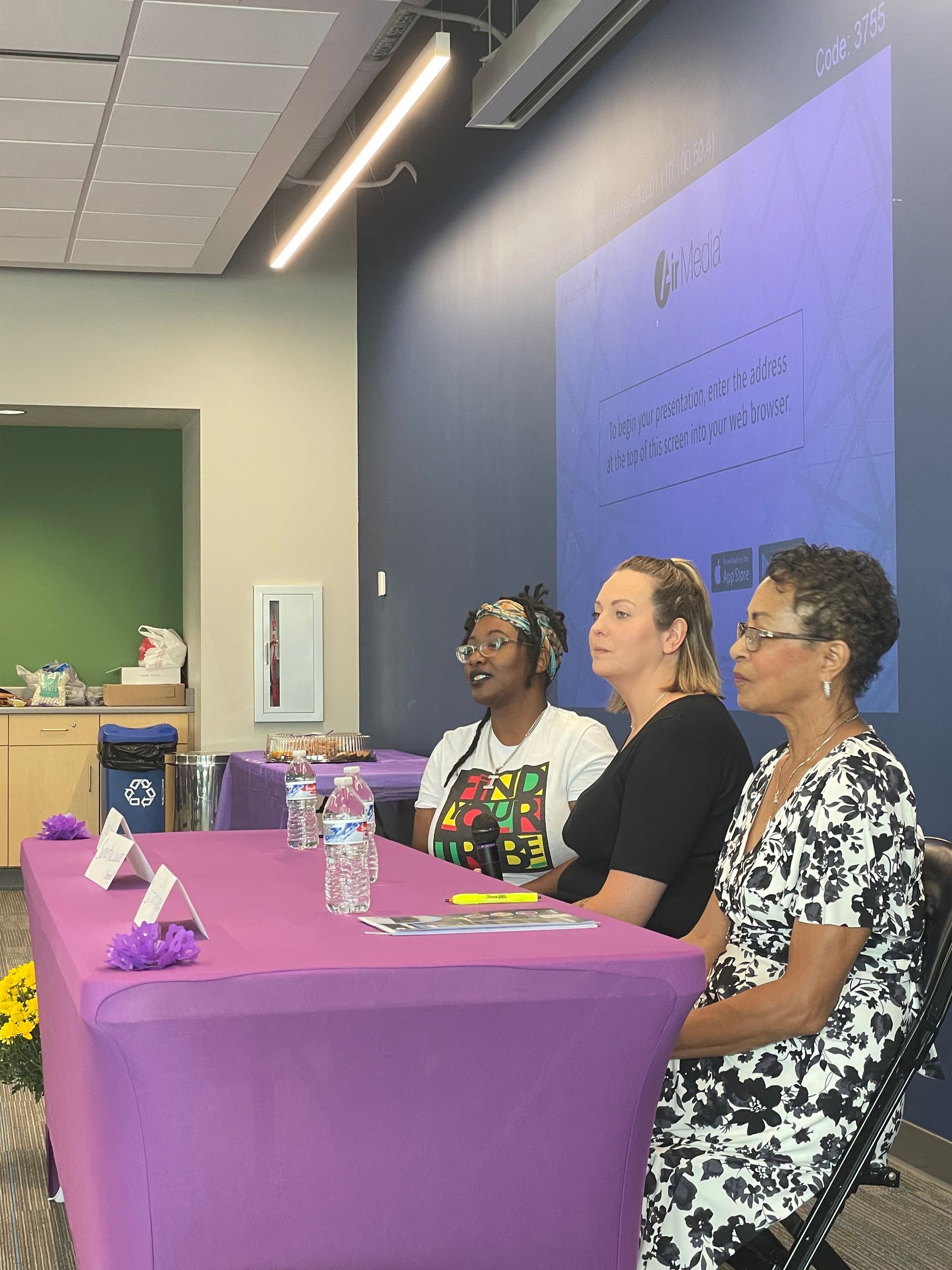 Three women are sitting at purple tables in a room.