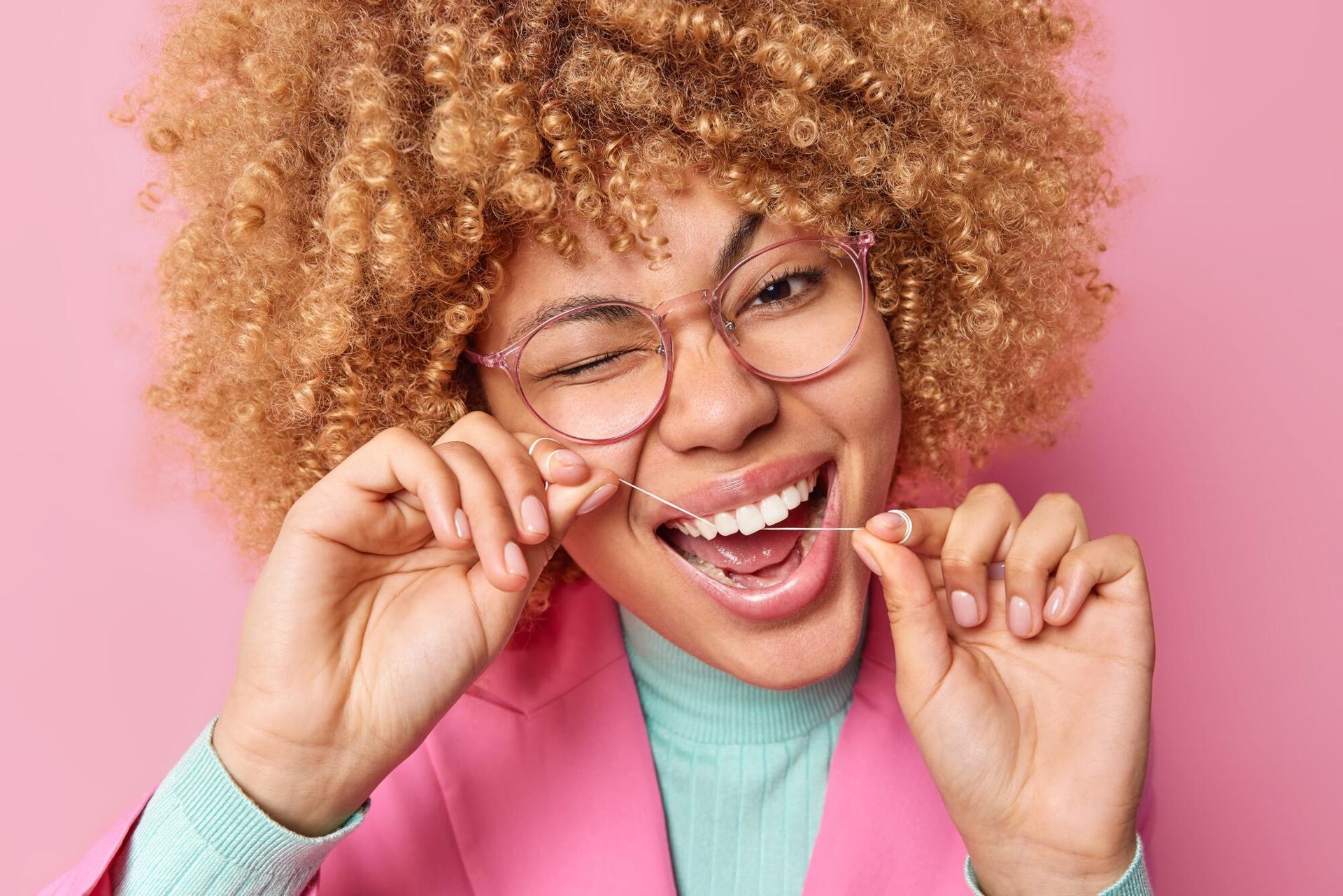 Woman with curly hair flossing teeth, winking, wearing glasses and pink blazer.