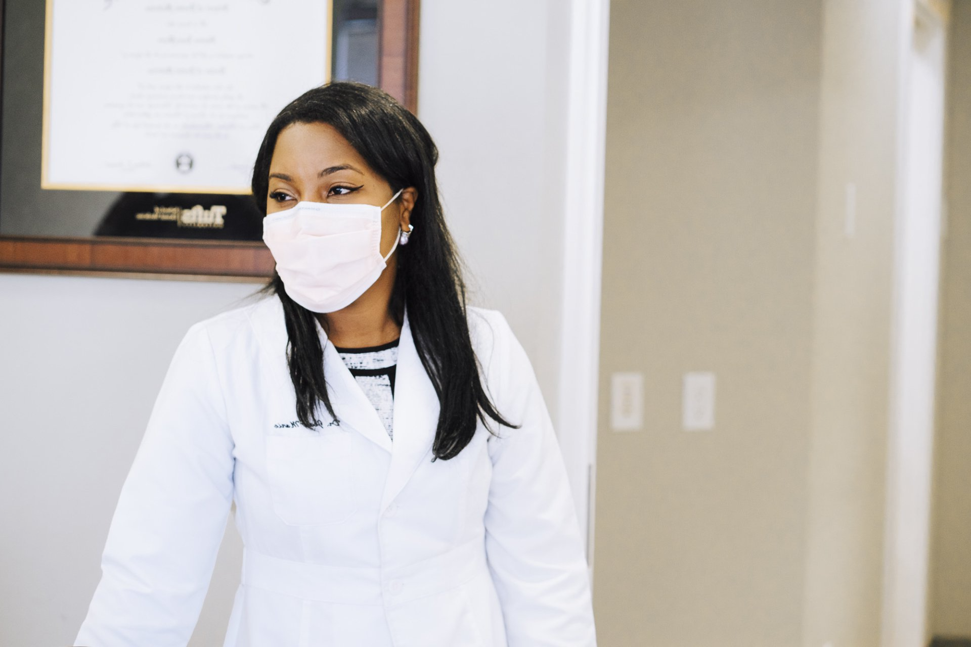 A female doctor wearing a mask and a white coat is standing in a hallway.