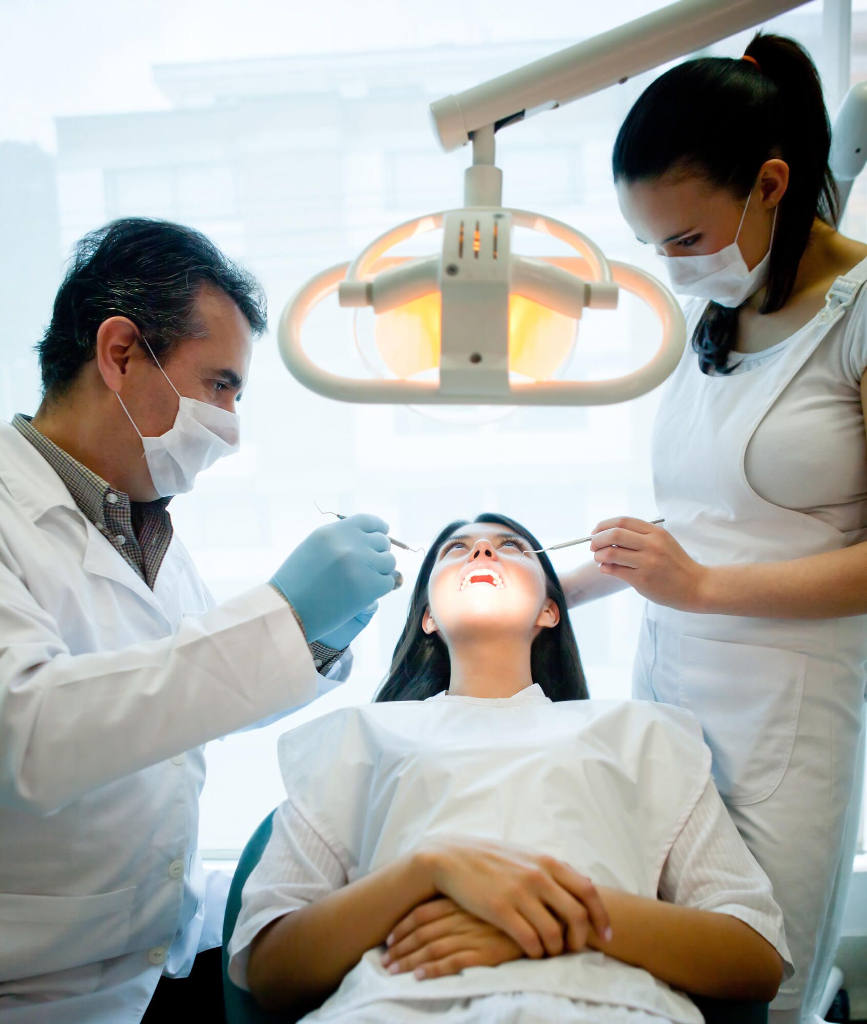 A dentist and assistant wearing surgical masks work on a patient lying in a dental chair under bright overhead lights.
