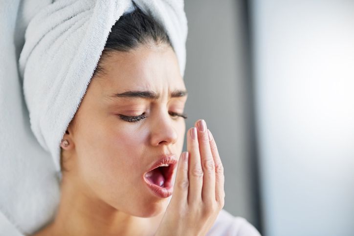 Woman with towel on head checking her breath, grimacing.