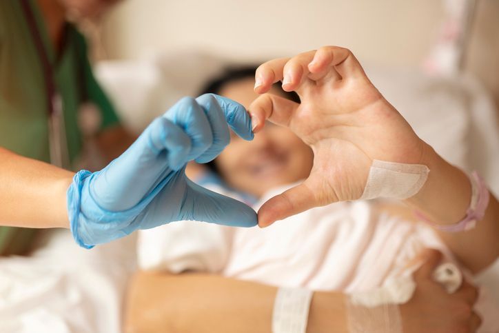 Nurse in blue gloves forming a heart shape over a patient in a hospital bed.