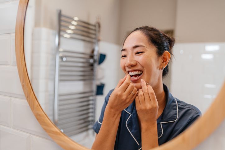 Woman in bathroom, smiling, using fingers to check teeth near a mirror.